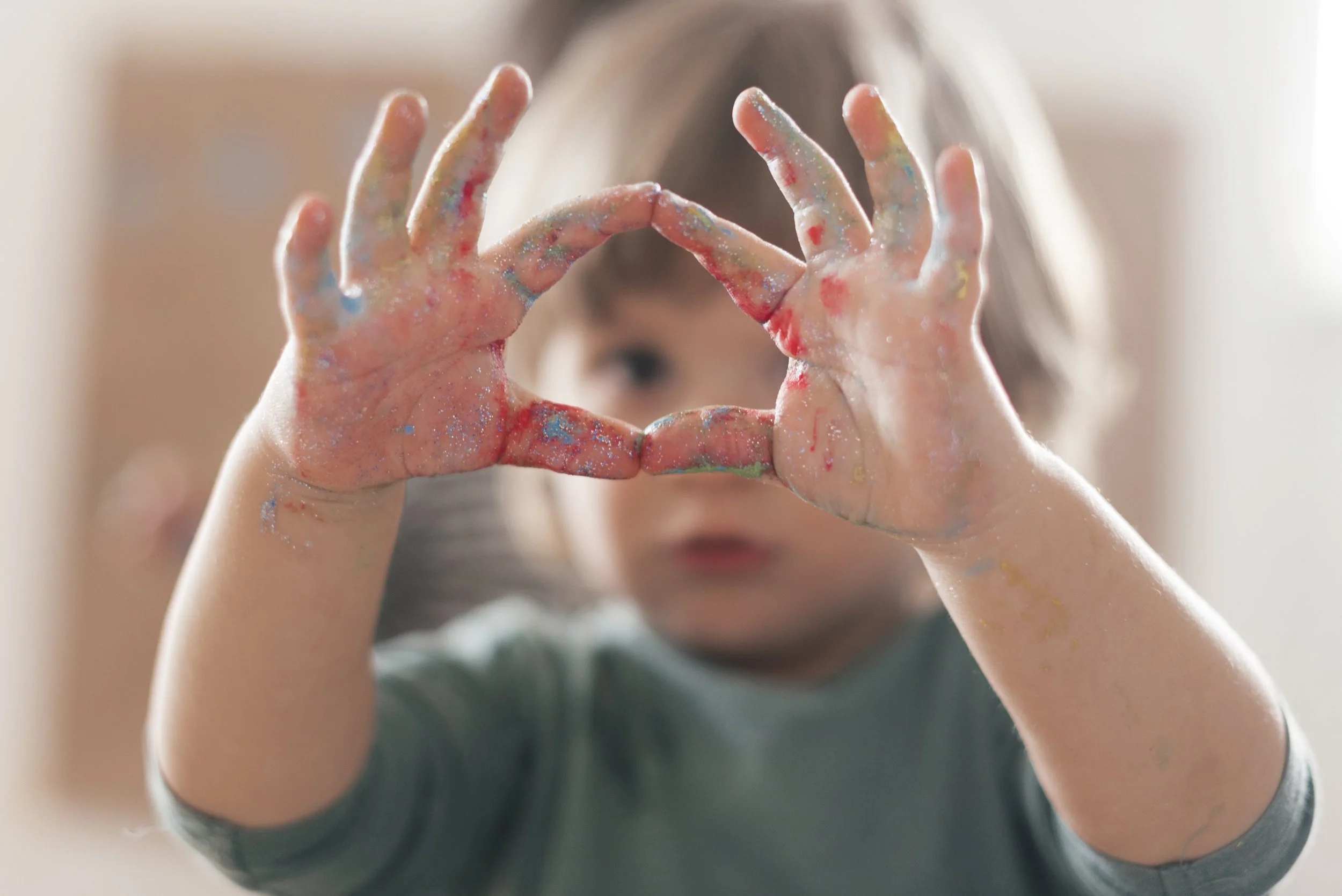 Child making a heart shape with hands covered in colorful paint.