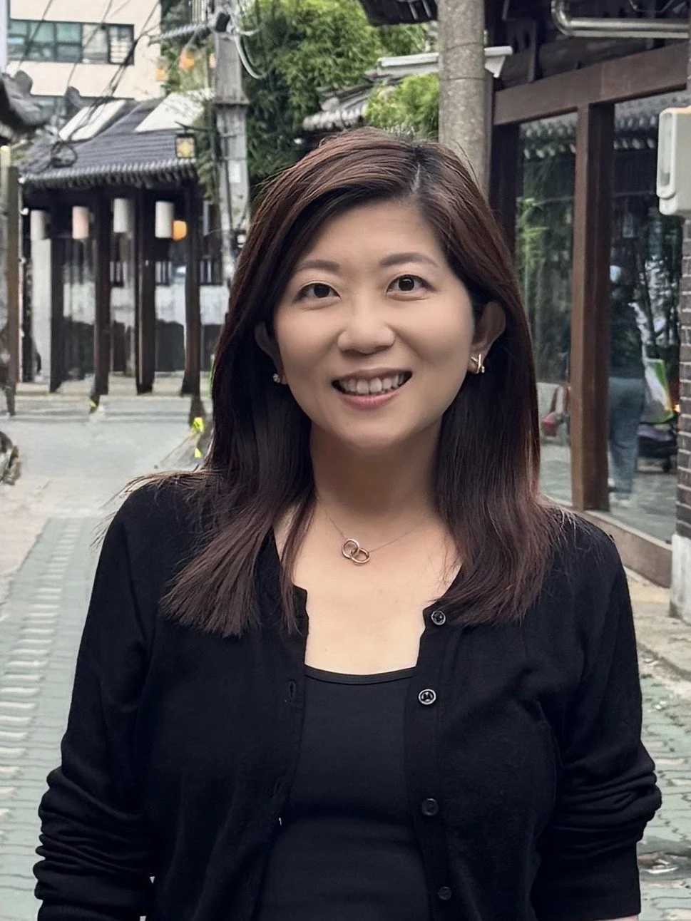 A woman with shoulder-length brown hair, wearing a black top and black cardigan, smiling outdoors in an urban setting with buildings and trees in the background.