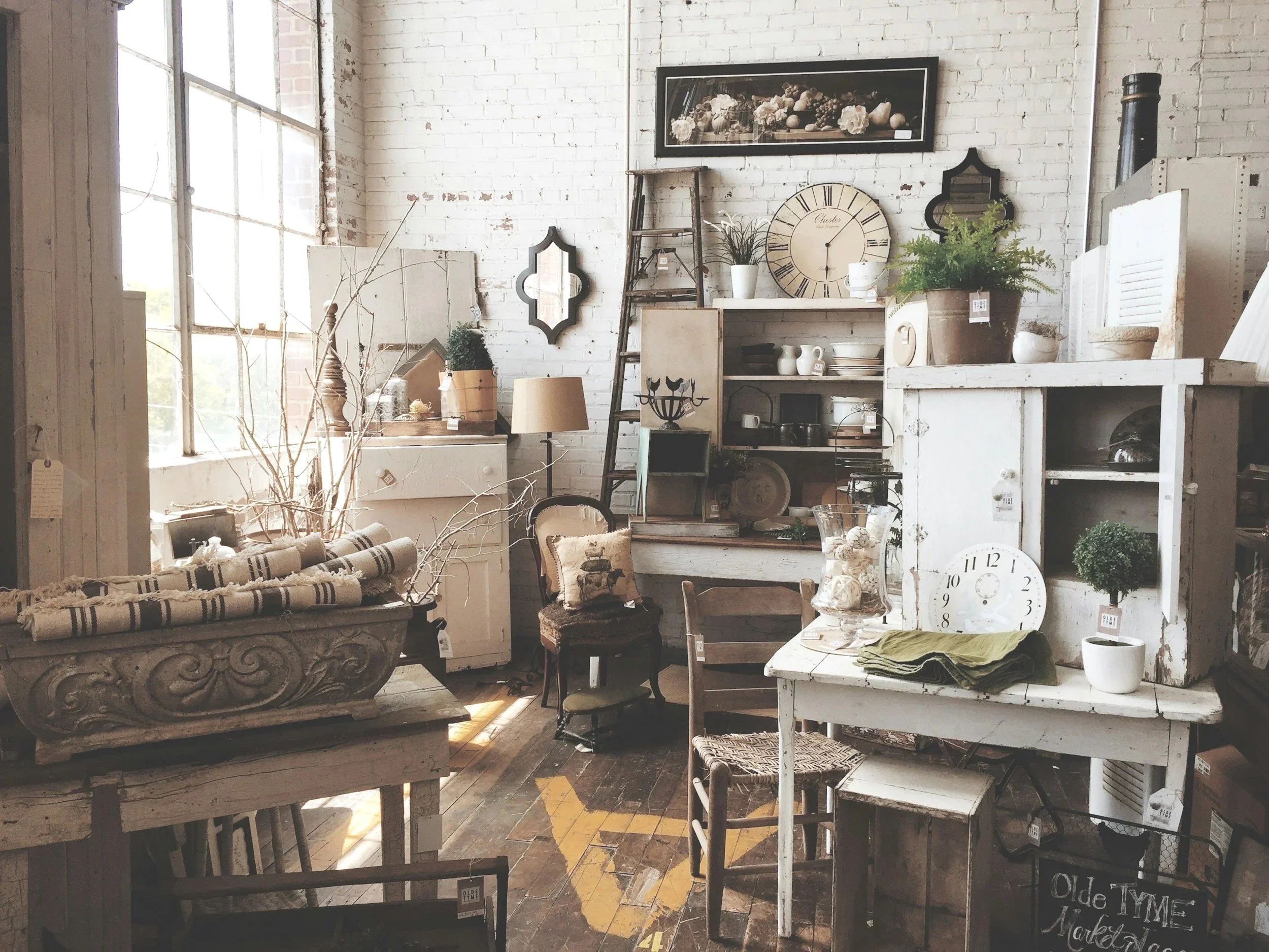 Interior of a rustic vintage store with white brick walls, large windows, and assorted antique furniture and decorative items such as clocks, plants, shelves, and chairs.