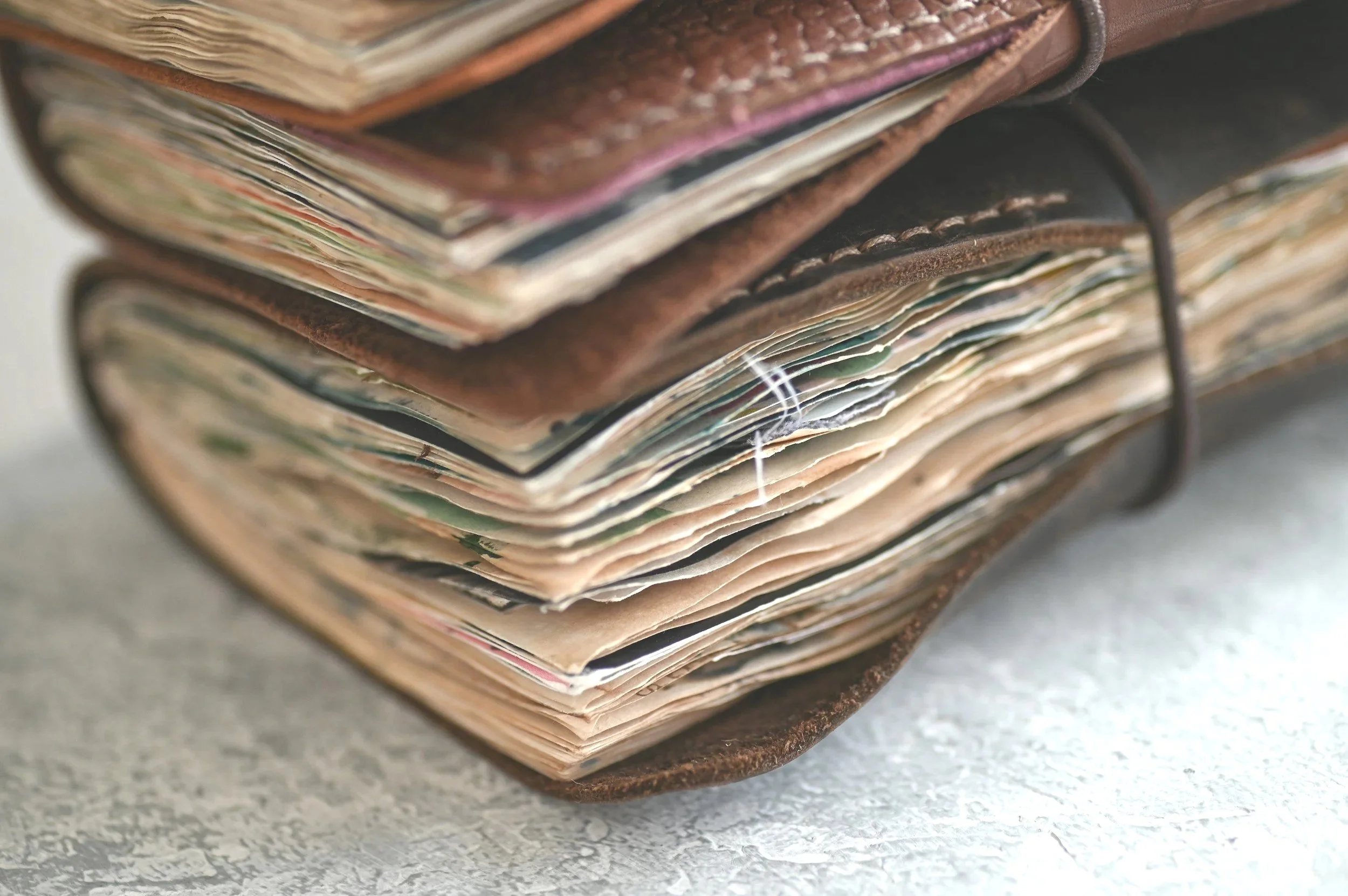 A close-up of an old, worn leather-bound scrapbook or journal filled with yellowed and torn pages resting on a textured surface.
