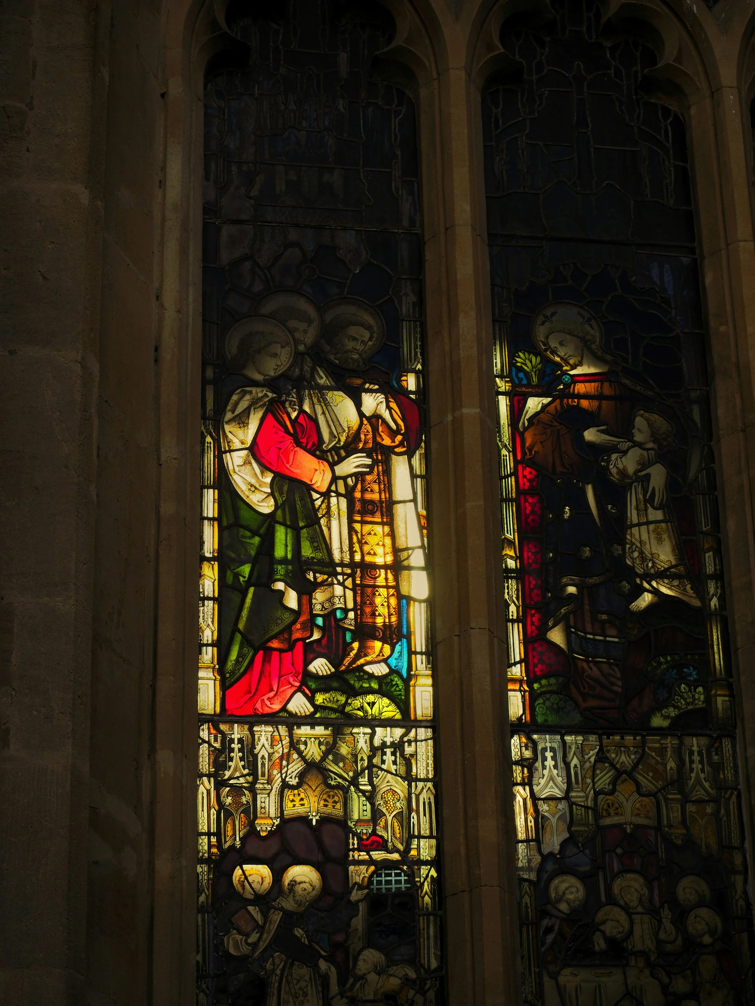 Two stained glass windows inside a church depicting religious figures, with one showing Jesus holding a cross and a woman, and the other showing Jesus with a child, both surrounded by various symbols and heraldry.