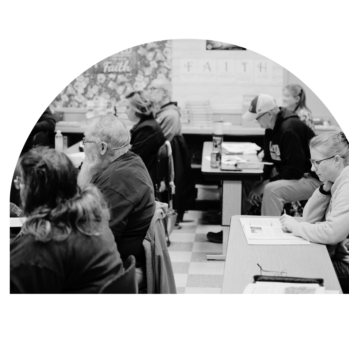 Black and white photo of people sitting in a classroom or seminar, reading and listening attentively, with books and papers on the tables, and the word 'FAITH' visible on a wall