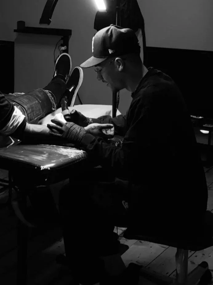 A man getting a tattoo on his forearm by a tattoo artist in a tattoo studio, with the man sitting on a chair and the artist kneeling beside him.