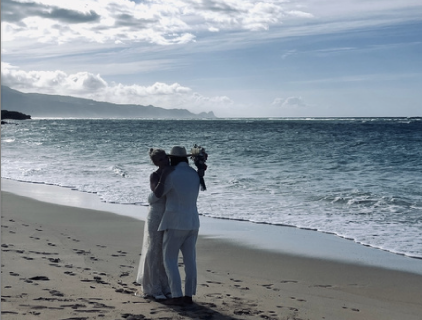 A couple dressed in white wedding attire embracing on a beach with the ocean and cloudy sky in the background.