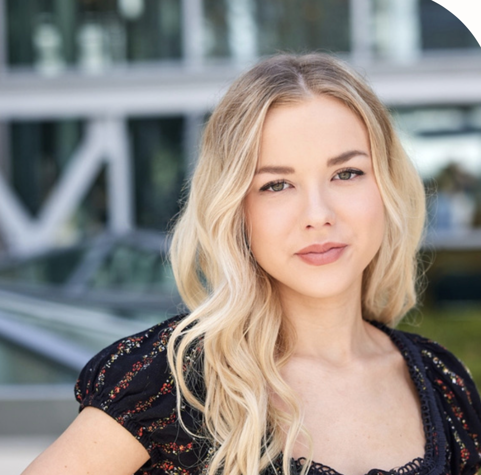 A young woman with blonde hair and light makeup standing outdoors with a blurred structure in the background.