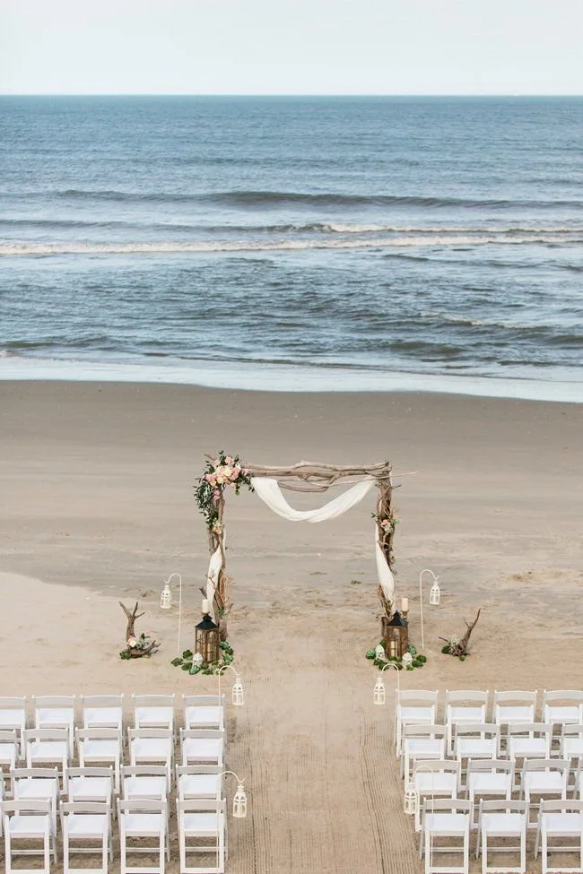 Wedding setup on a beach featuring a wooden arch decorated with flowers and fabric, surrounded by white chairs, lanterns, and ocean waves in the background.