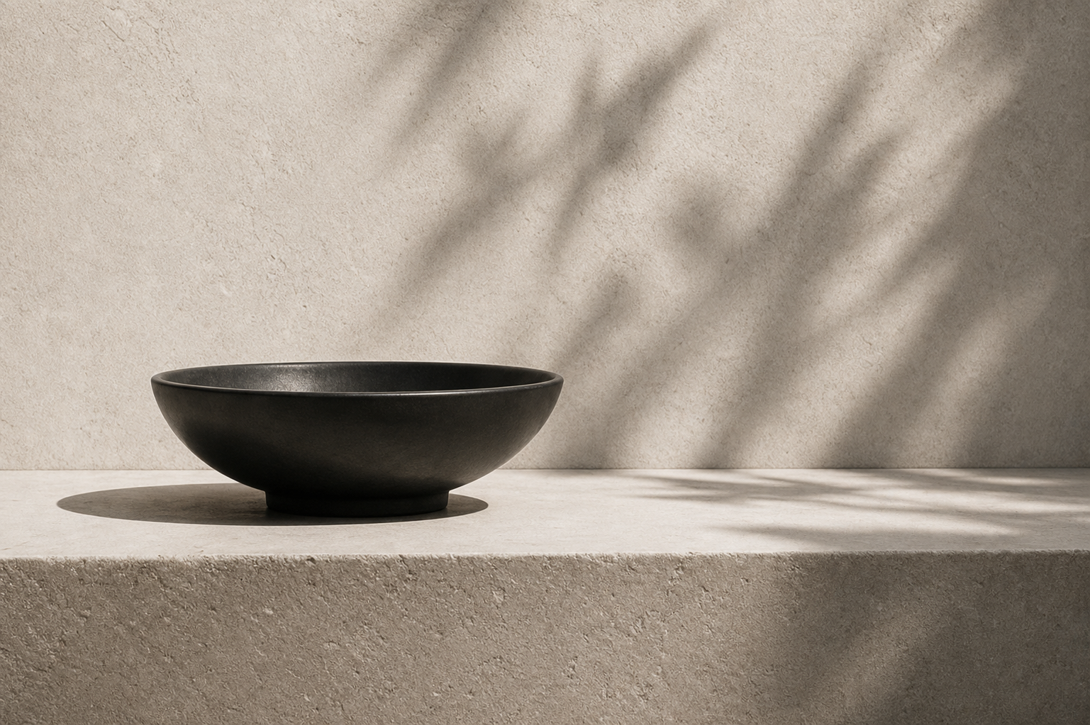 A black ceramic bowl on a beige textured surface with shadows cast by a plant or tree on a beige wall.