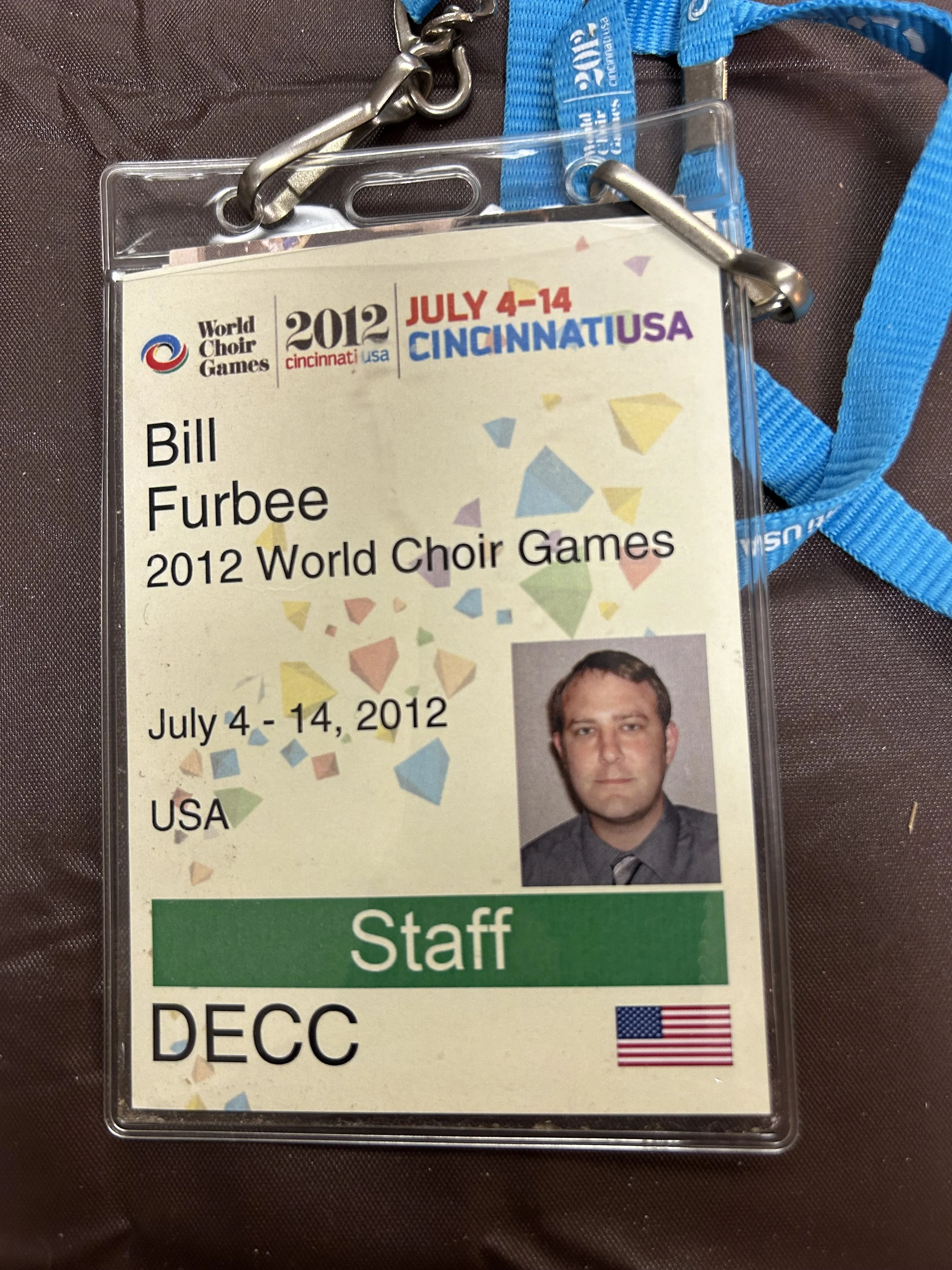 A staff badge for the 2012 World Choir Games in Cincinnati, USA, worn by Bill Furbee. The badge includes a photo of Furbee, the event dates July 4-14, 2012, and the American flag.