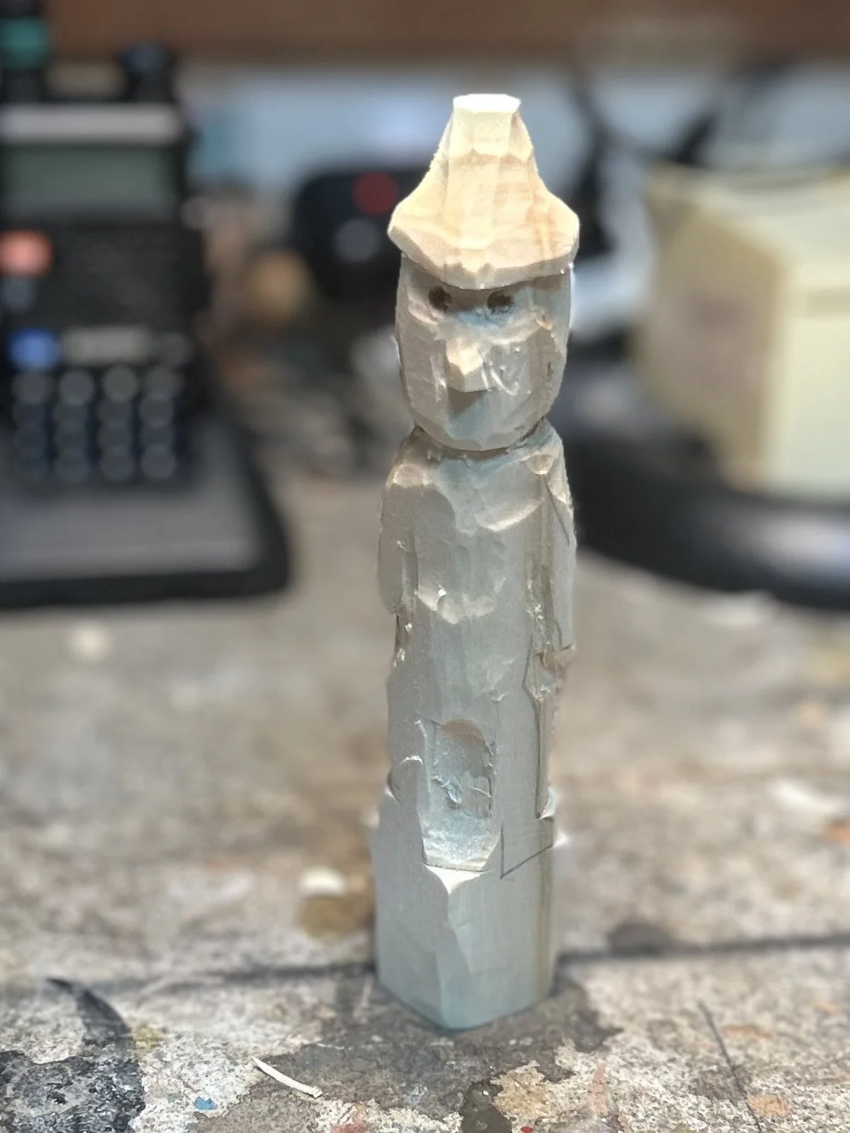 Wooden carving of a figure with a pointed hat, standing on a workbench with woodworking tools in the blurred background.