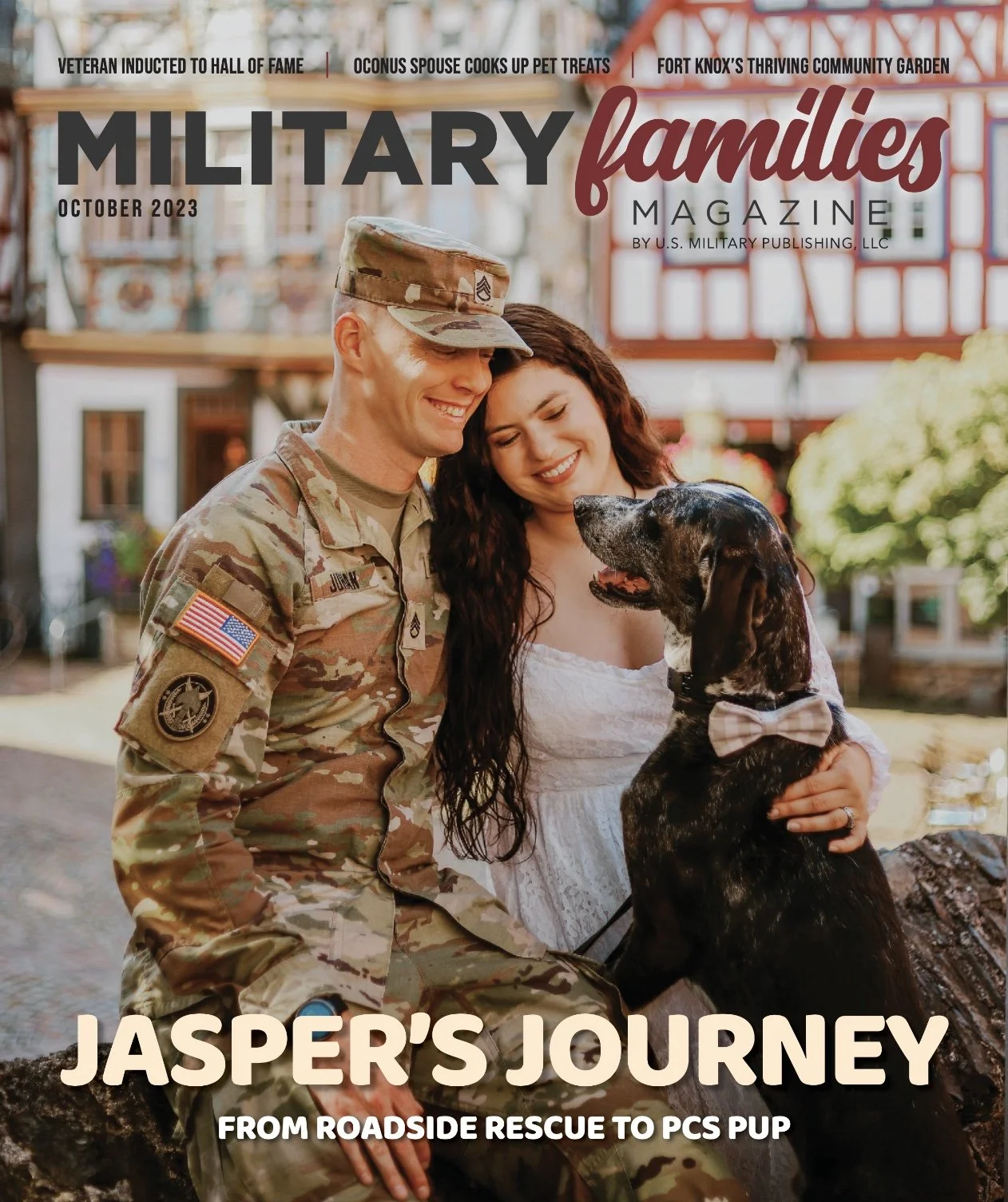 A soldier in camouflage uniform sitting on a log, smiling with a woman in a white dress, and a black dog with a bowtie sitting beside them, outdoors in front of a building with colorful decorations.