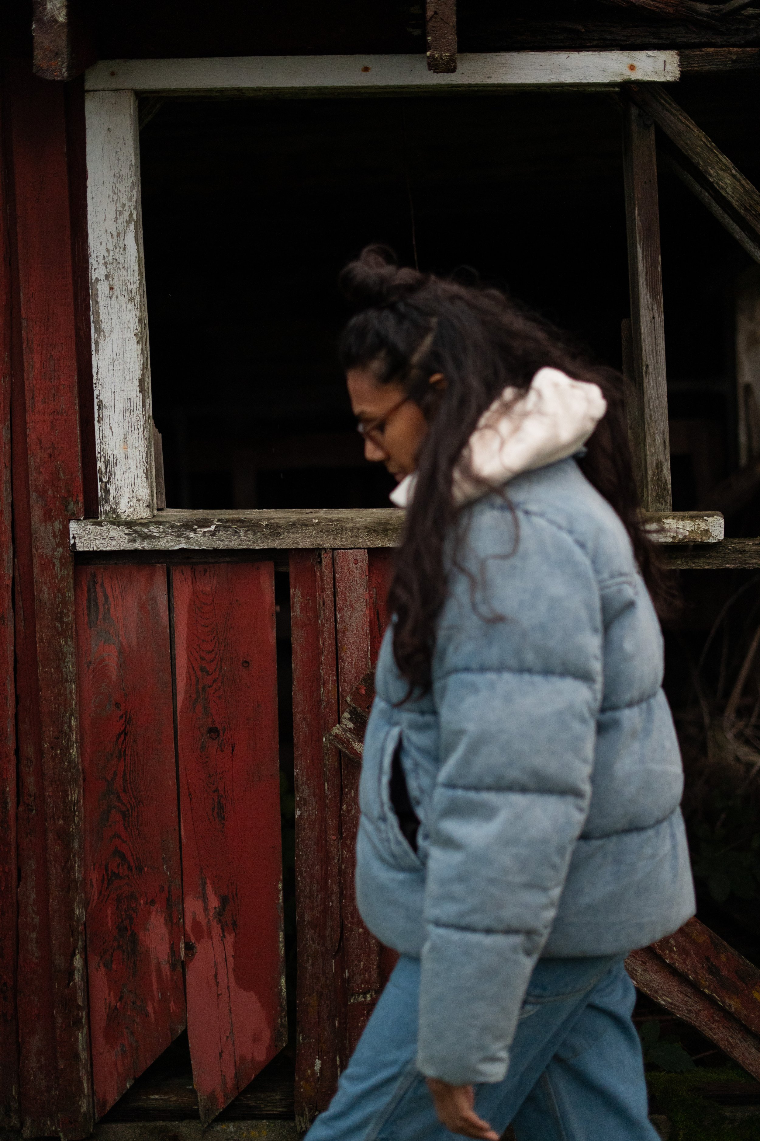 Desiree Dawson walking past an old red barn with a window framed in white paint wearing a blue faux denim puffer jacket and hair in a half up half down with a bun wearing glasses with brown frames her out of focus with her head looking down