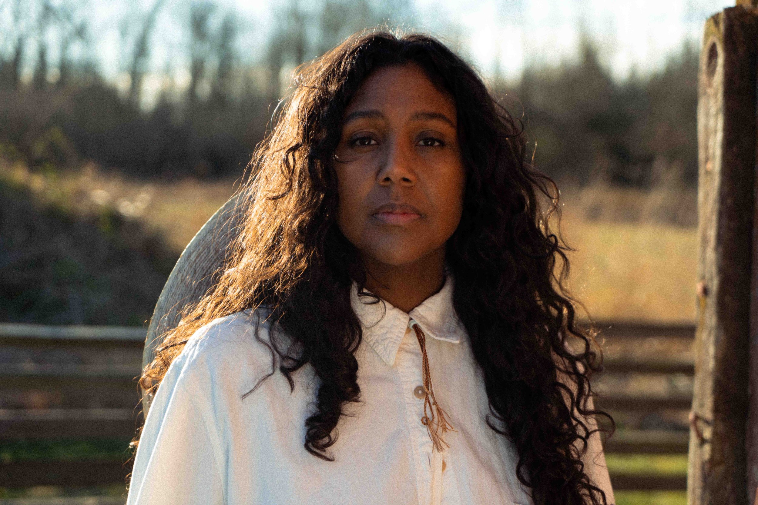 Desiree Dawson looking into the camera with warm sunlight on the side of her curly hair her cowboy hat around her neck and sitting on her back wearing a white blouse and a field and forest behind her