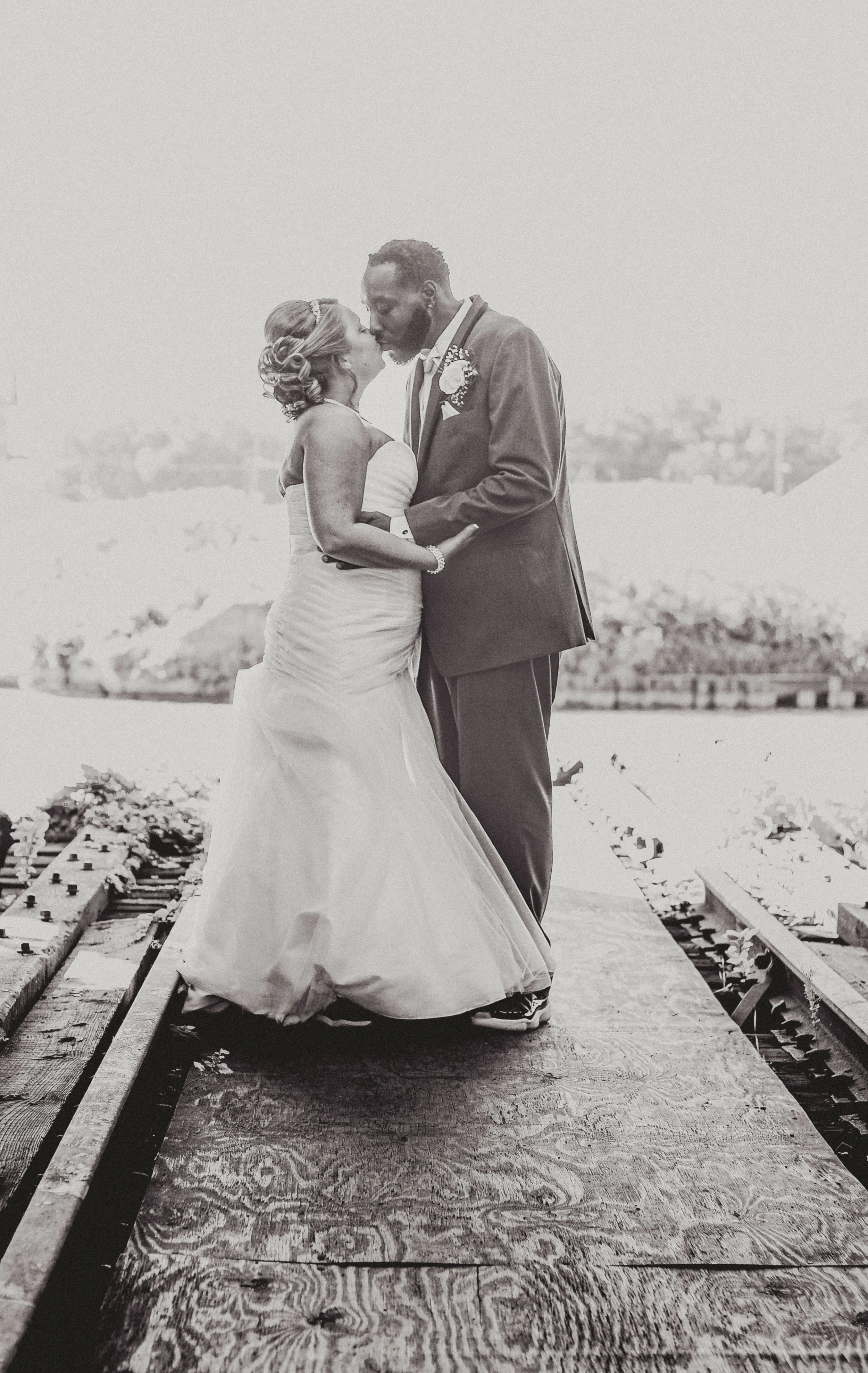 Black and white photo of a bride and groom kissing on a dock over water. The bride is in a strapless wedding dress, and the groom is in a suit with a boutonniere.