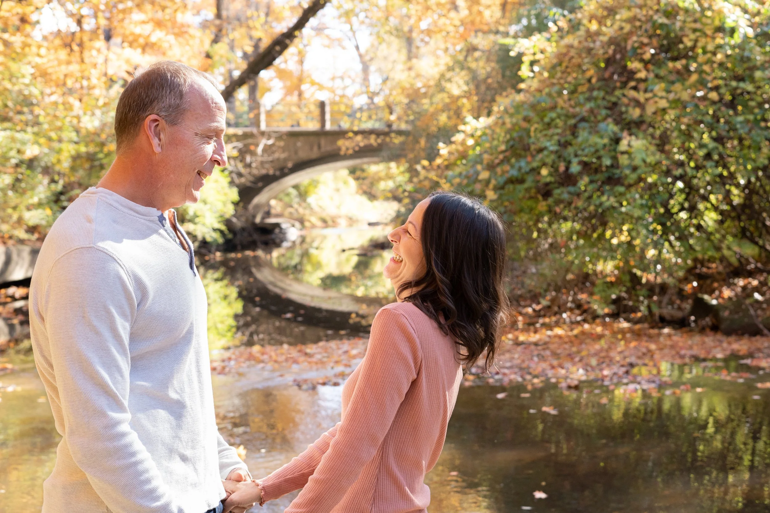 A man and woman holding hands, smiling, and looking at each other in an outdoor autumn setting near a creek with colorful fall foliage.