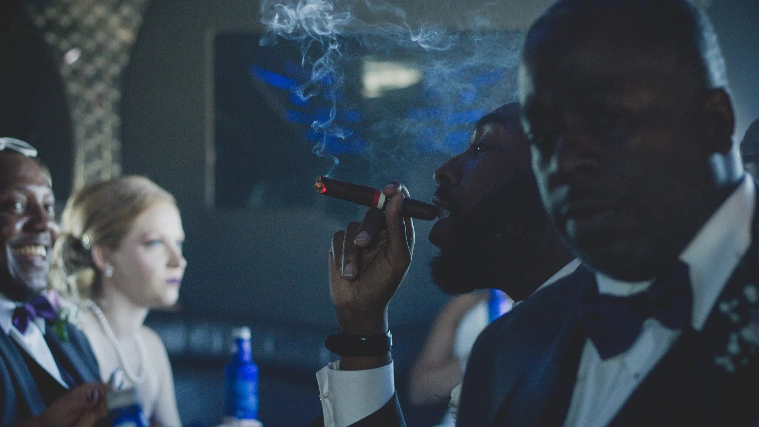 A man in a tuxedo smoking a cigar at a formal event with other elegantly dressed people around him.