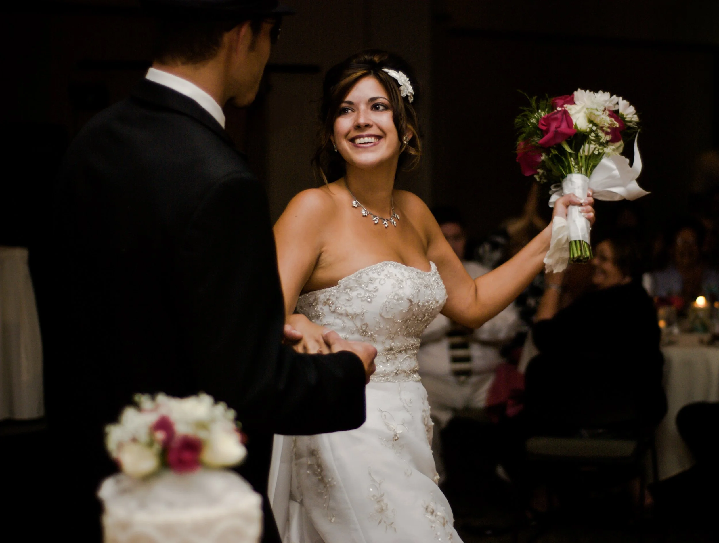 A bride in a strapless wedding gown holding a bouquet of pink and white flowers, smiling at a groom in a black tuxedo during a wedding reception.