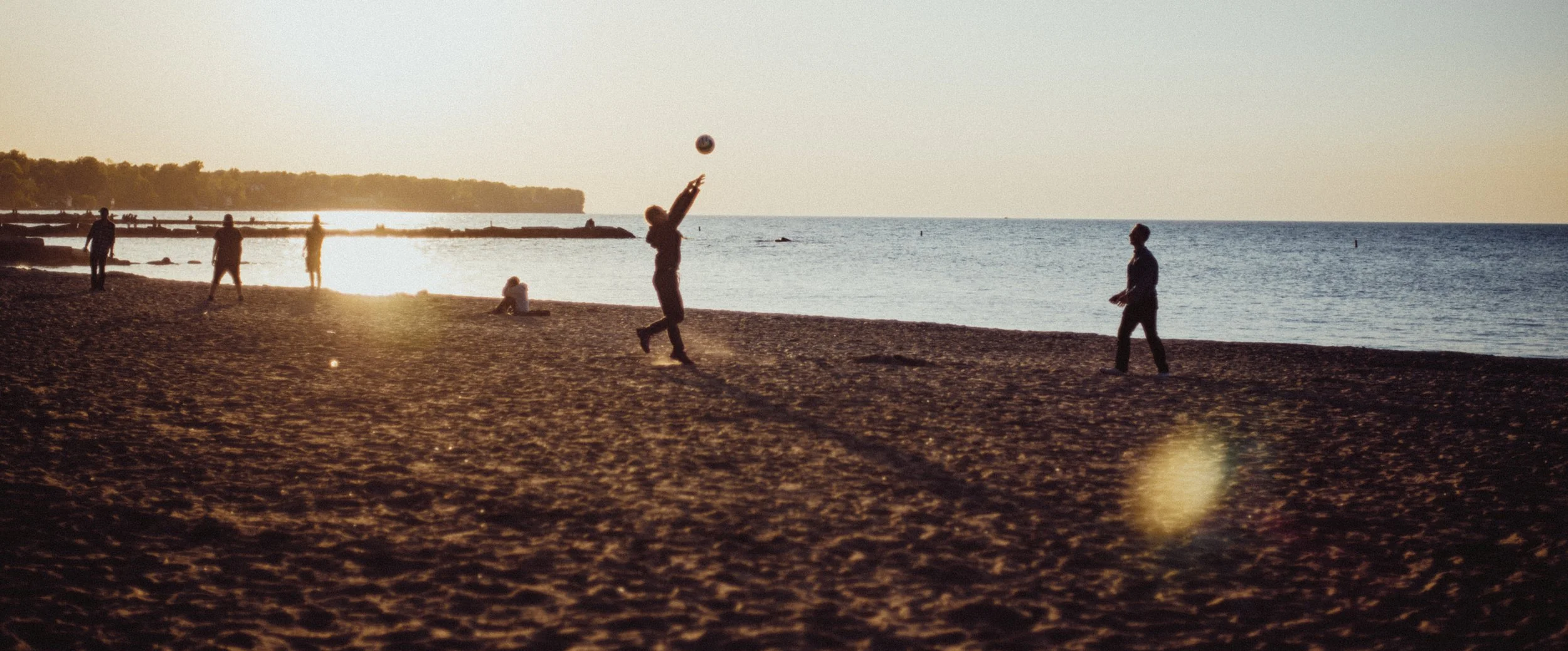 People playing beach volleyball at sunset by the water.