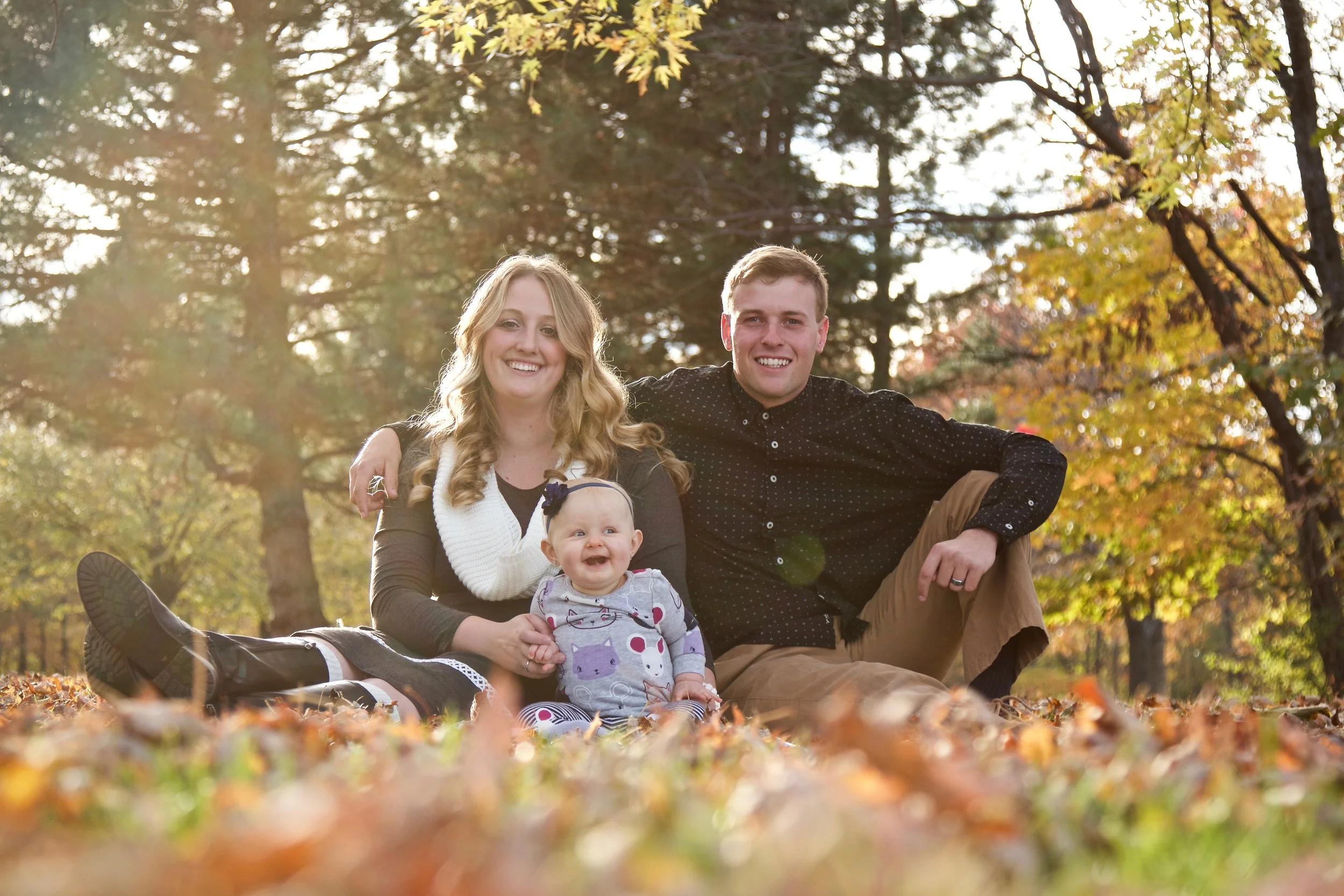 Family of three sitting on fallen autumn leaves in a park with trees and sunlight in the background.