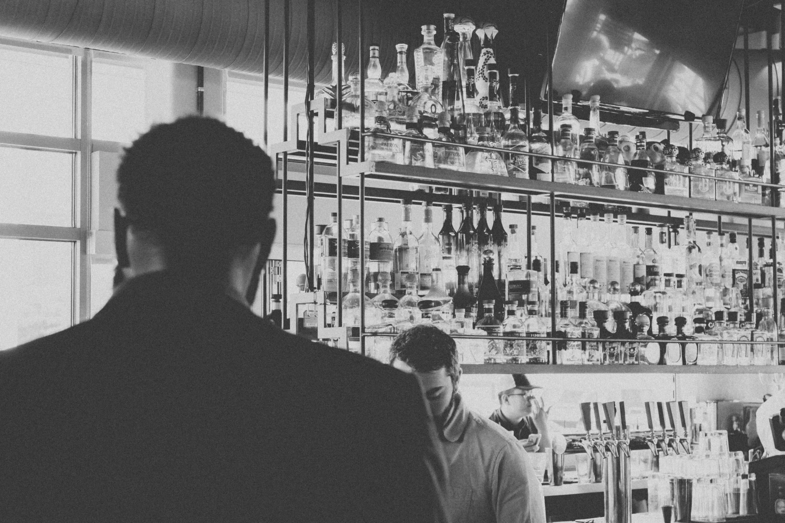 Black and white photo of a bar with shelves stocked with bottles, seen from behind a person with short curly hair. Two bartenders are visible, one looking down and the other with a hand near his face, in front of the bar counter.