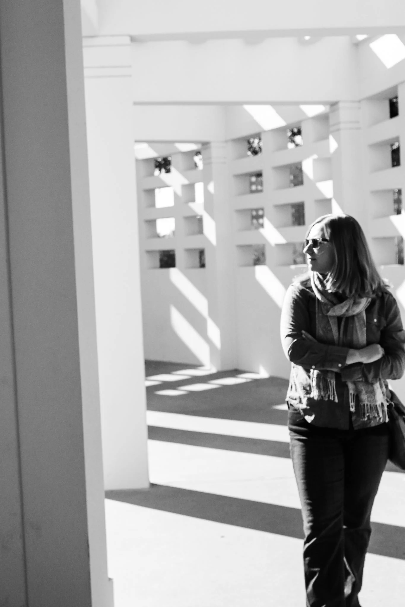A woman with glasses and a scarf stands outdoors with her arms crossed, looking to the side, with sunlight creating shadows on a white building in the background.