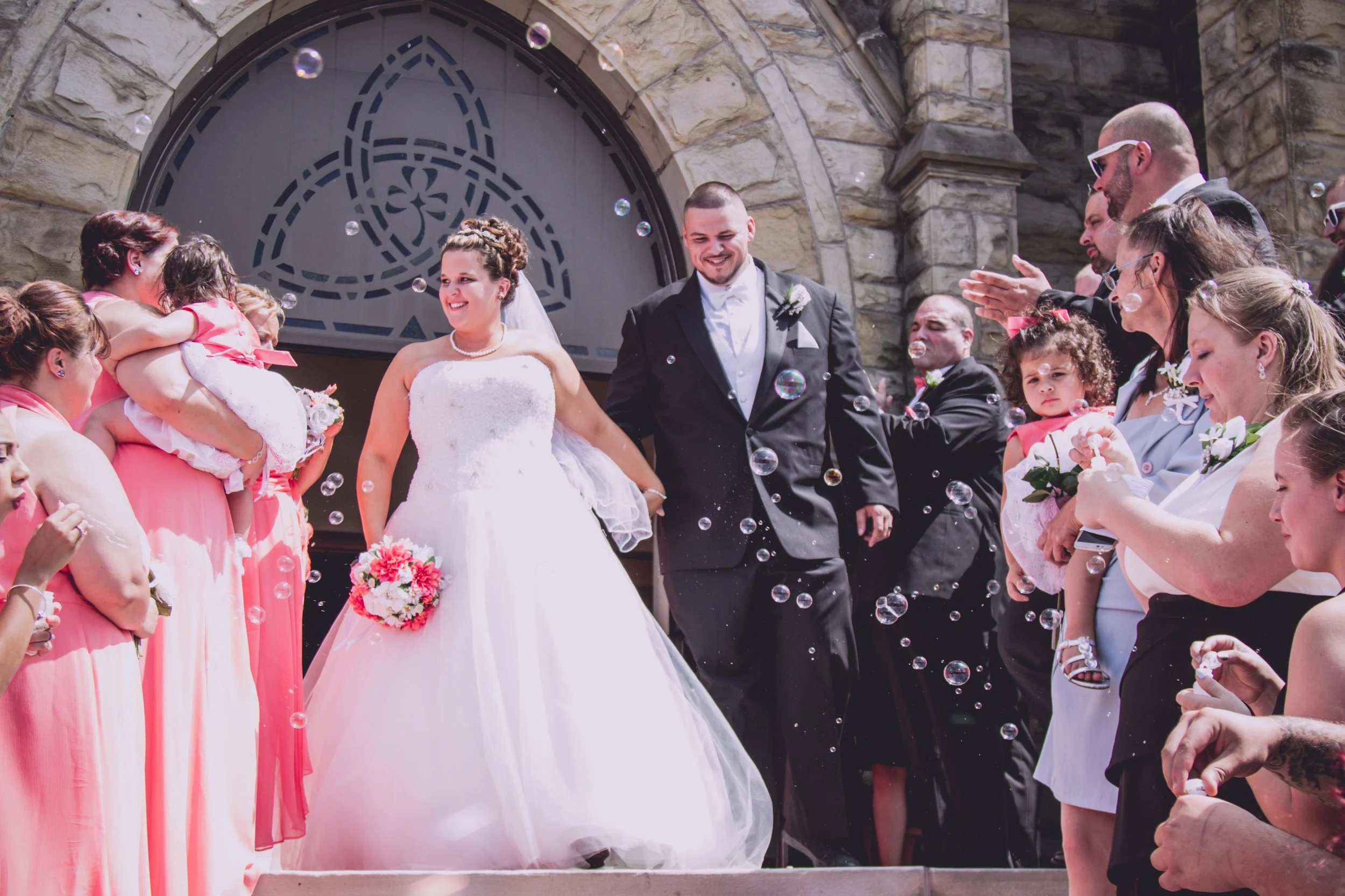A bride and groom holding hands and smiling as they walk down the steps outside a stone church, surrounded by friends and family celebrating with bubbles.