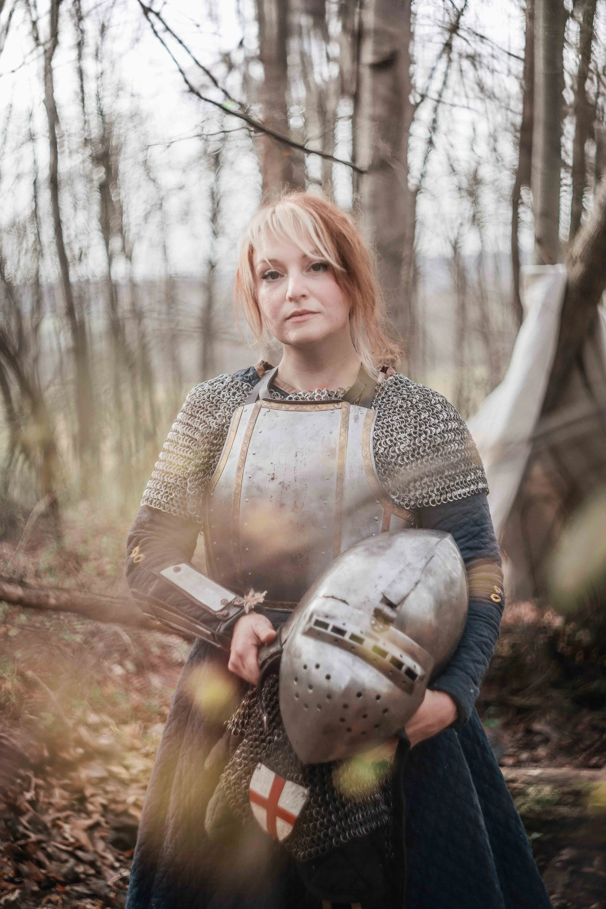 Woman wearing medieval armor holding a helmet in a wooded area during daytime.