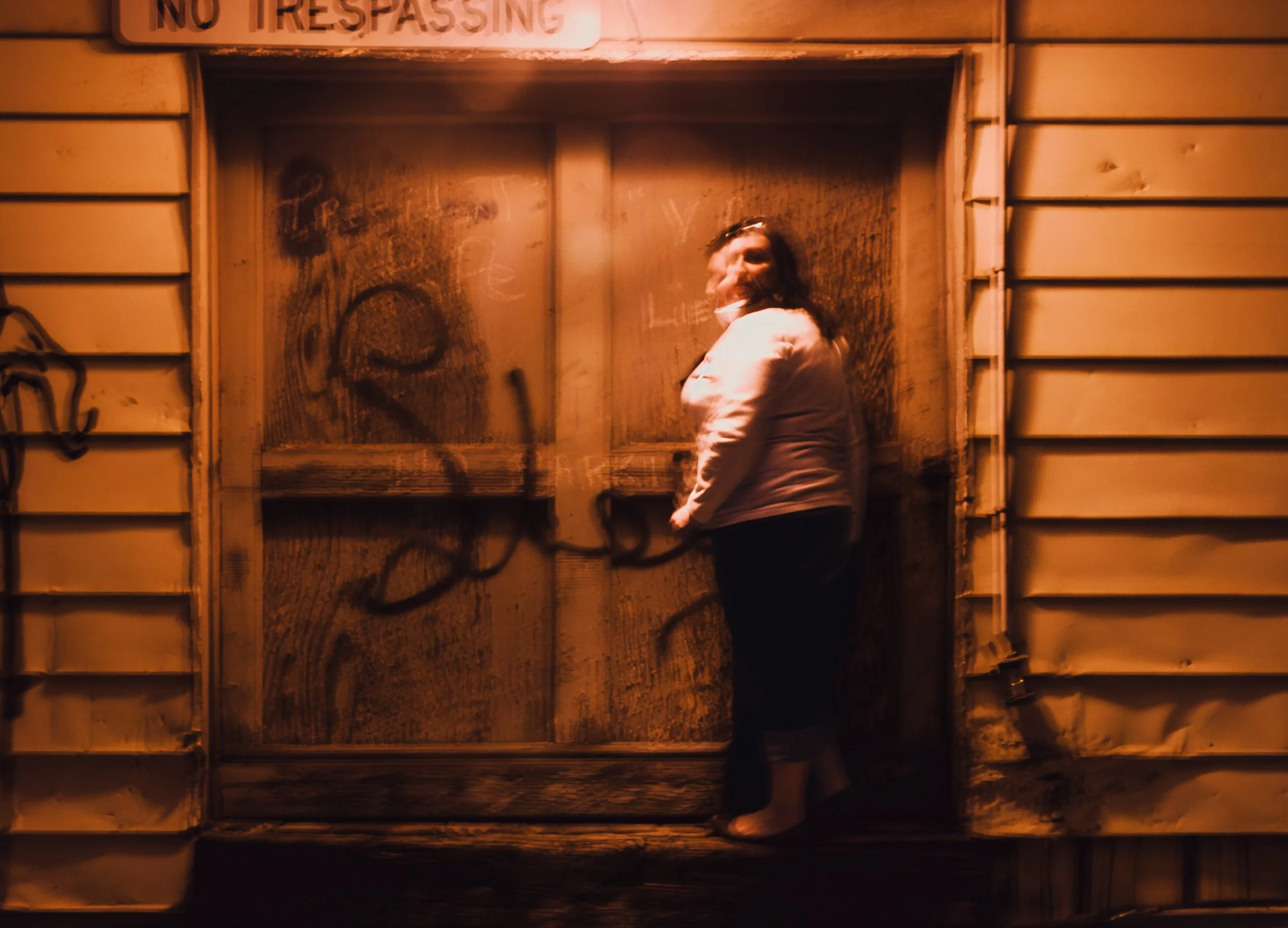A woman standing in front of a worn wooden door with graffiti, illuminated by warm orange light. The door has a 'No Trespassing' sign on top.