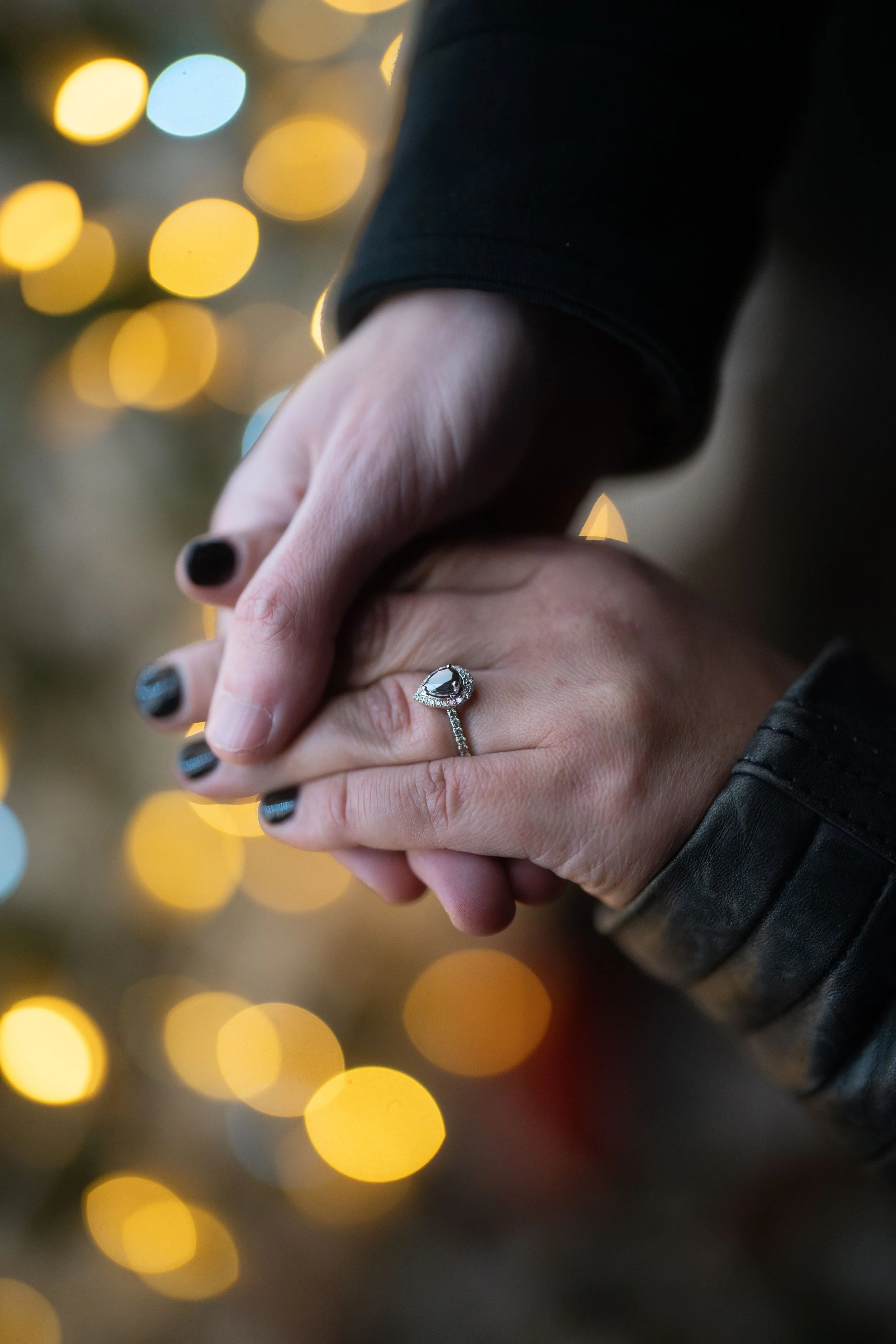 Close-up of two hands holding each other, one with an engagement ring, against a background of blurred festive lights.