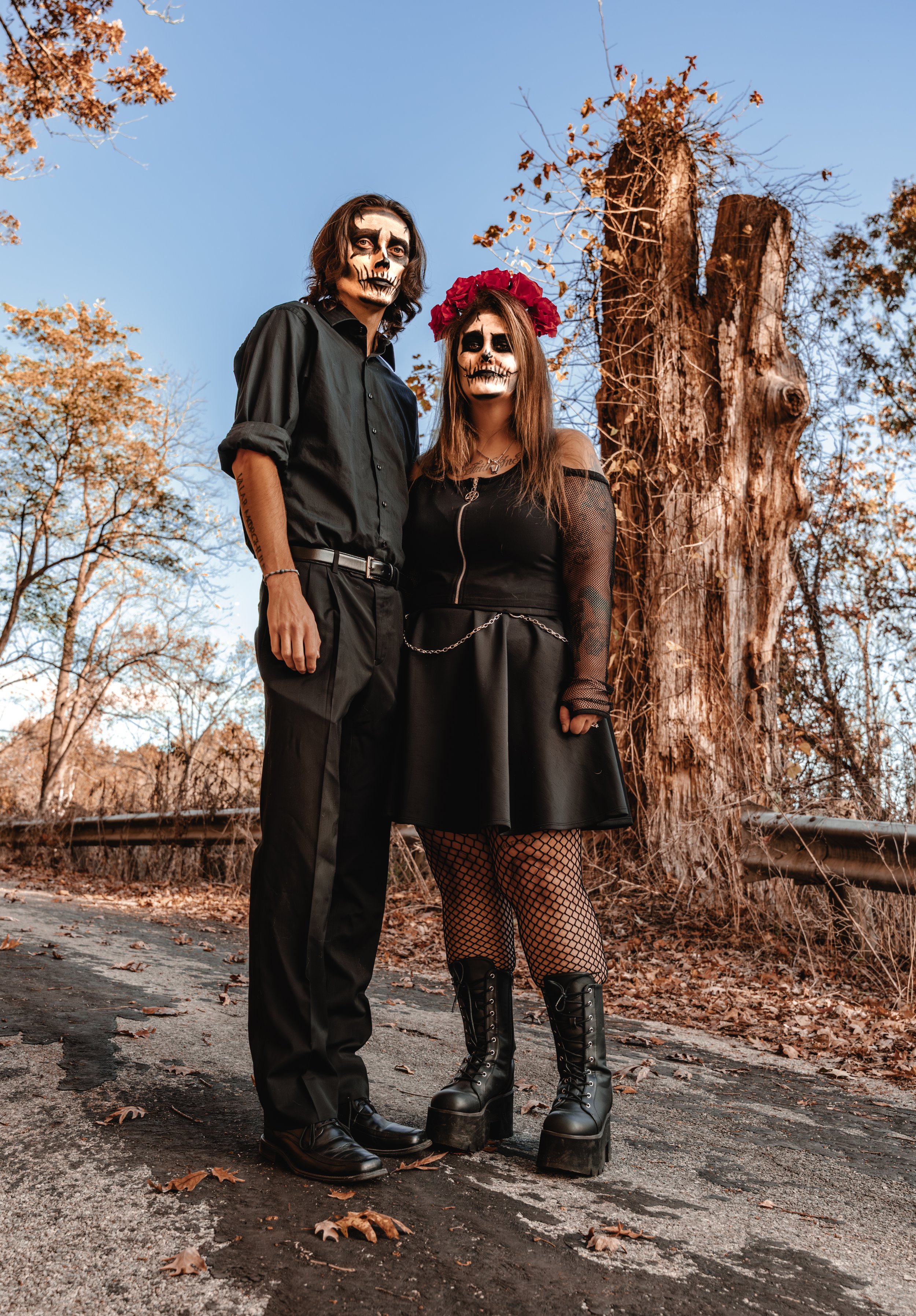 Two people dressed in skeleton makeup and black clothing standing on a dirt road with autumn trees and a large tree trunk behind them under a clear blue sky.