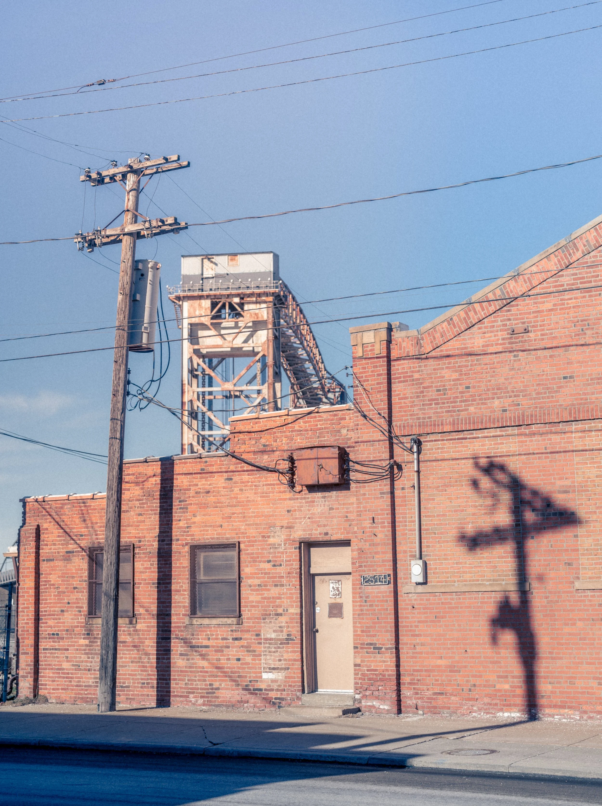 A brick building with three windows and a door, electrical wires, a utility pole, and a shadow of a utility pole on the wall, with a water tower in the background against a blue sky.