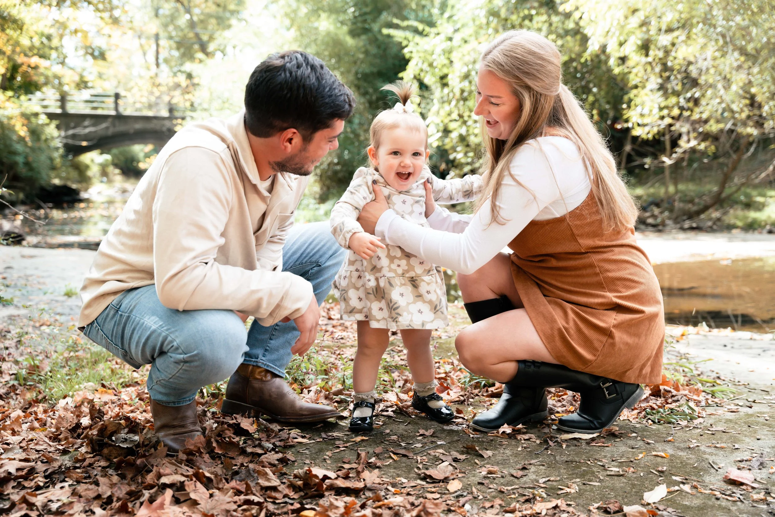 A happy family of three, including a man, woman, and a young girl, is outdoors by a river, with tall trees and a bridge in the background. The woman is kneeling and holding the girl's hands, while the man crouches nearby, all smiling and enjoying a m