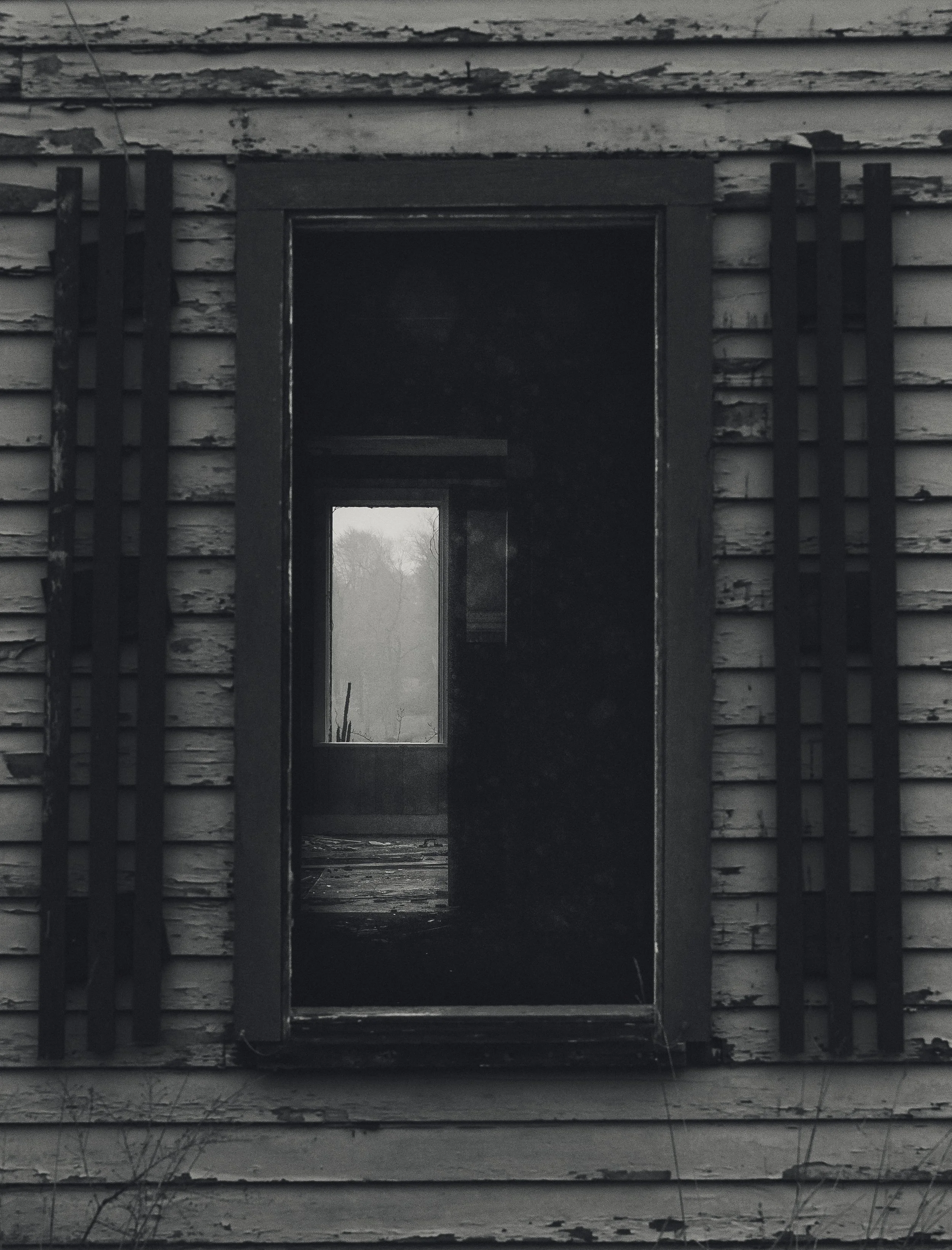 An old, weathered building with peeling paint, featuring a narrow doorway with no door and a small window at the back, framed by black decorative bars.