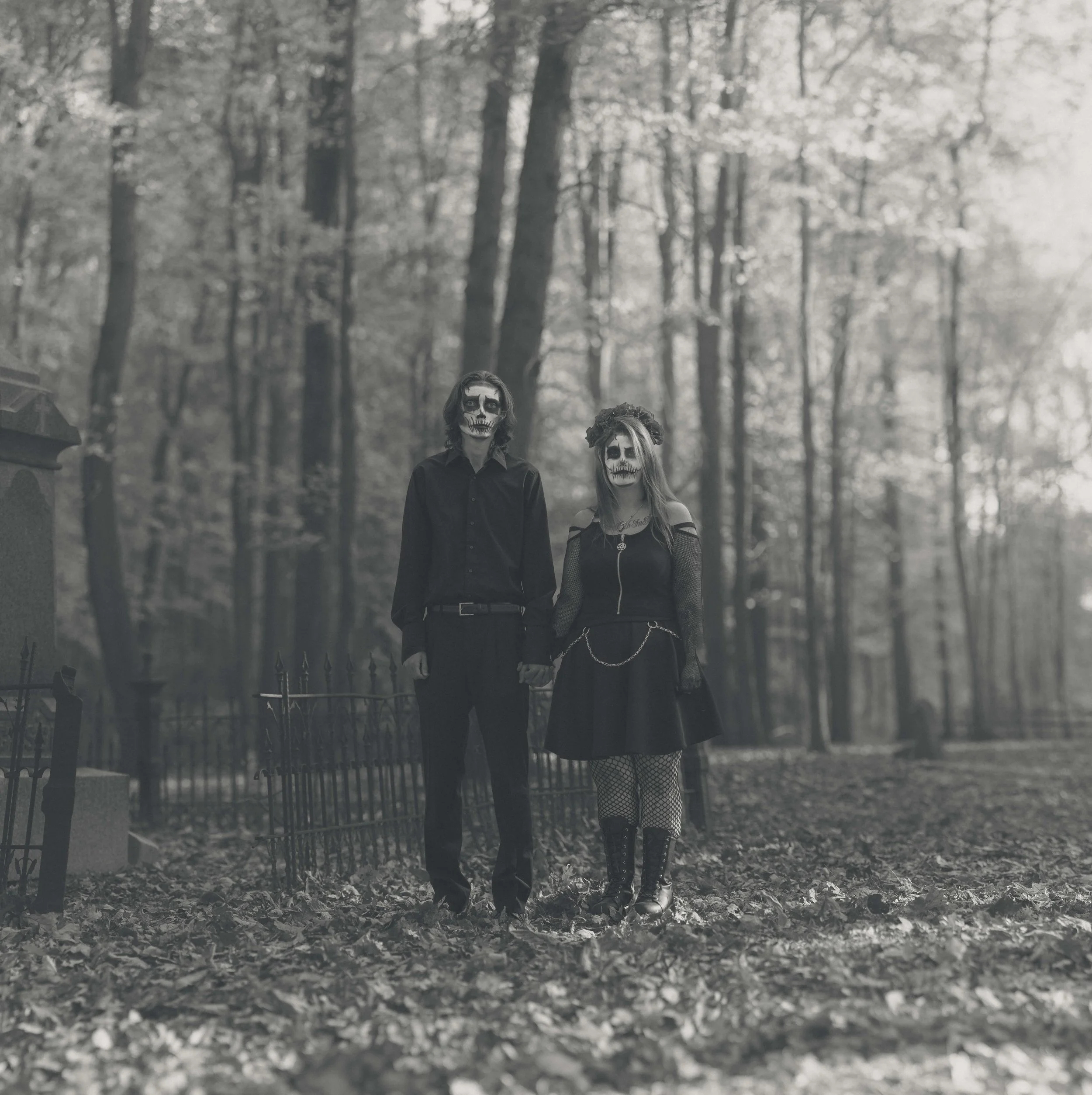 Couple dressed in gothic Halloween costumes with face paint resembling skulls, standing hand-in-hand in a forest with autumn leaves on the ground.
