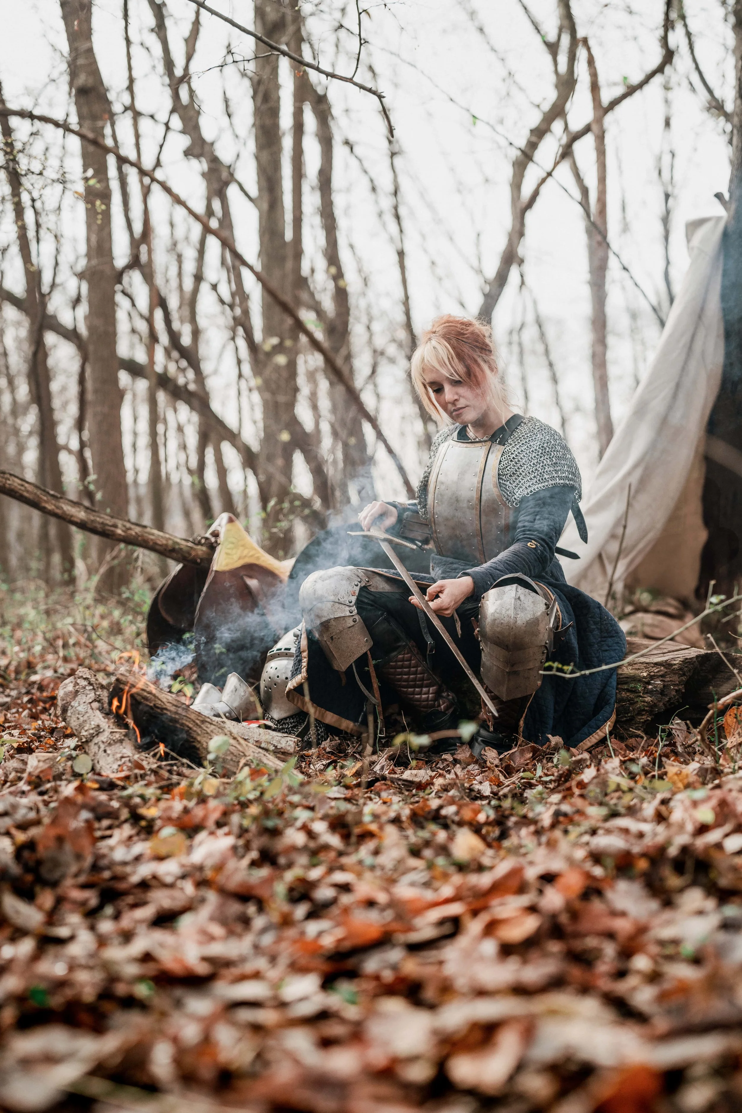 A person dressed in medieval armor sitting by a campfire in a wooded area during autumn.