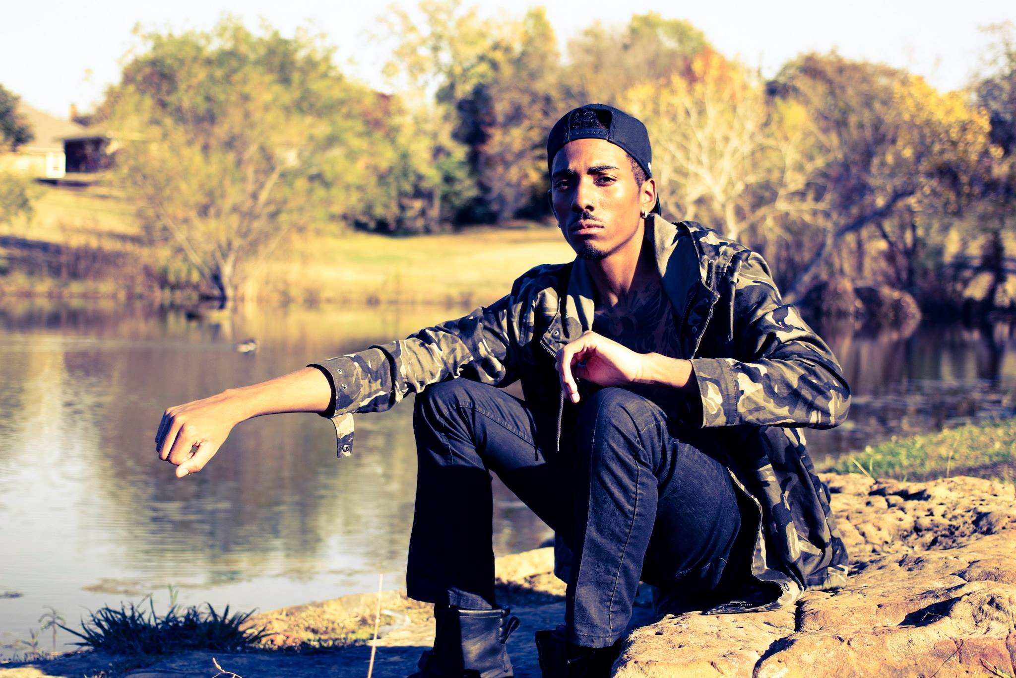 A young man with a serious expression, wearing a camouflage jacket and a black baseball cap backwards, squatting by a lake in a park with trees in autumn colors.
