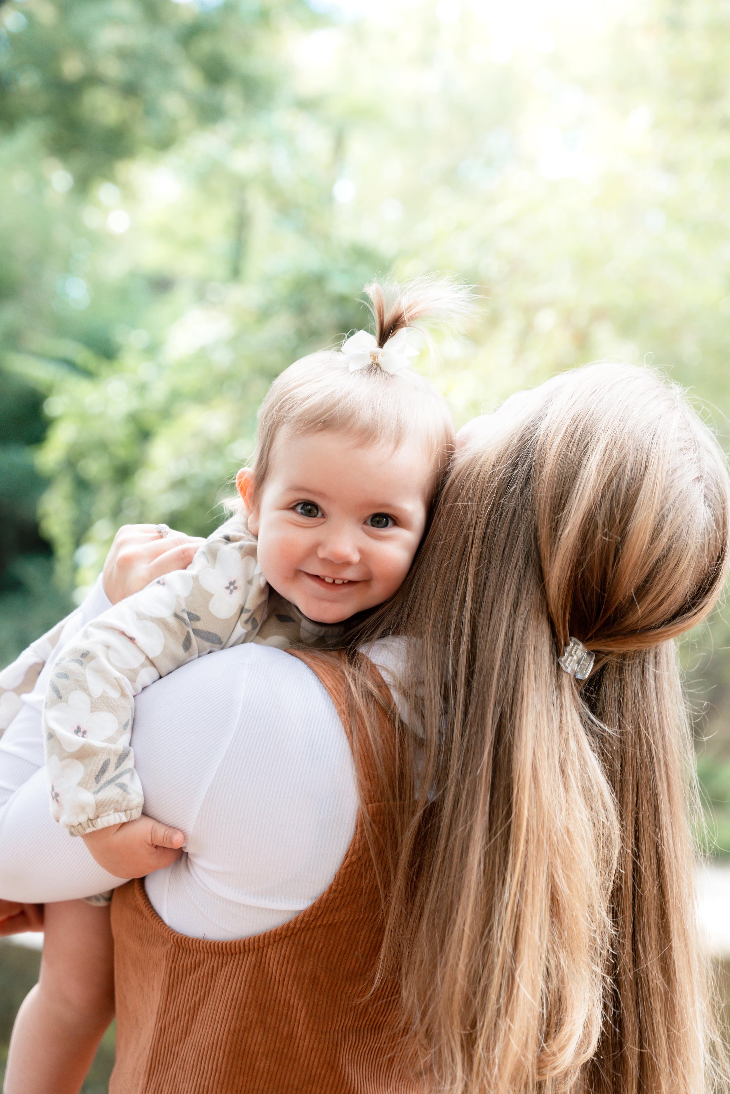 A young girl with a smiling face and a top knot hairstyle with a bow, being carried on an adult woman's back outdoors with a blurred green forest background.