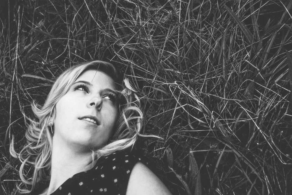 A woman lying on her back on grass or hay, looking upward with a thoughtful expression in a black-and-white photo.
