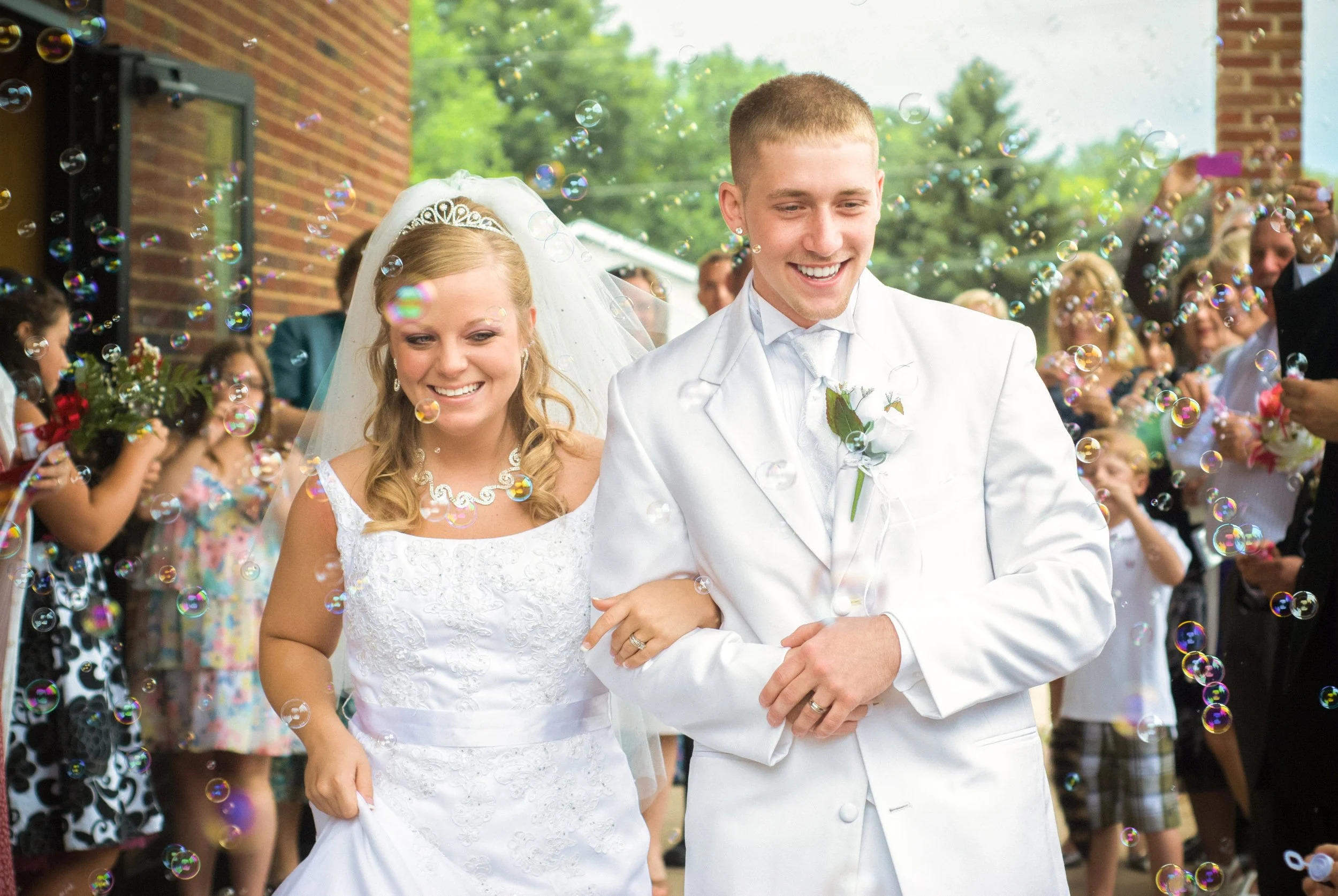 A smiling bride and groom walking arm-in-arm surrounded by family and friends celebrating their wedding outside, with bubbles floating around.
