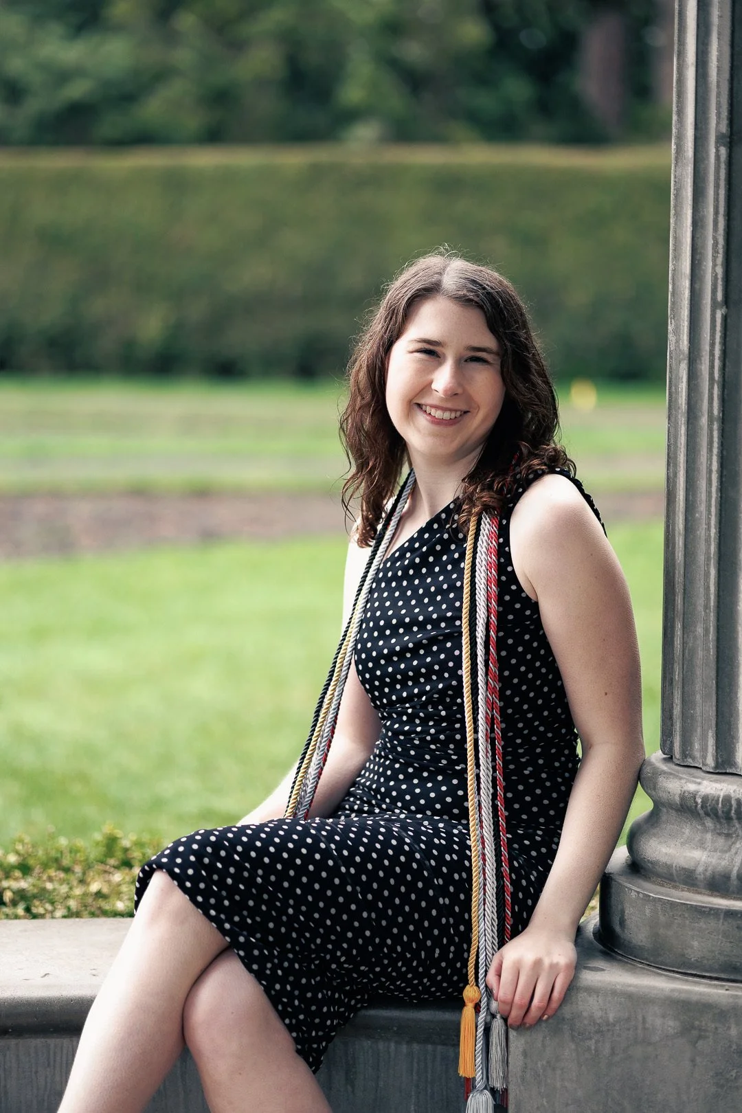 A young woman with curly brown hair sitting outdoors on a cement ledge, smiling, wearing a black polka-dot dress, with graduation cords around her neck, near a stone column, with a grassy field and trees in the background.