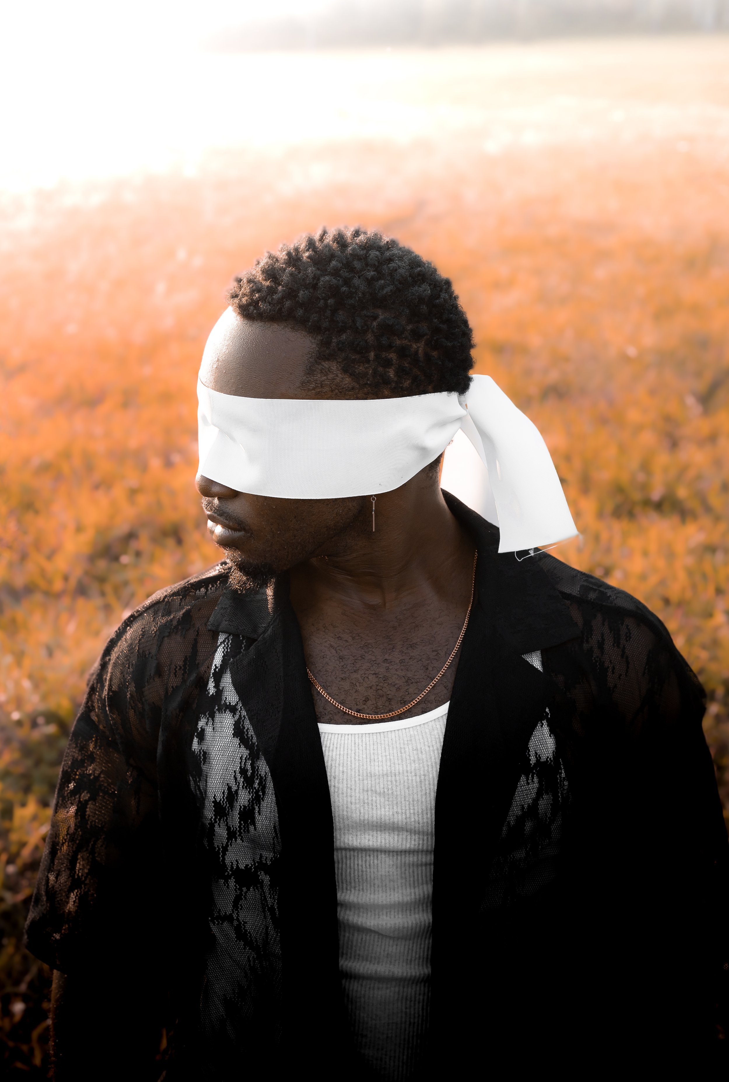 A man with curly hair and dark skin is blindfolded with a white cloth, standing outdoors on a field of orange and yellow vegetation during sunset.