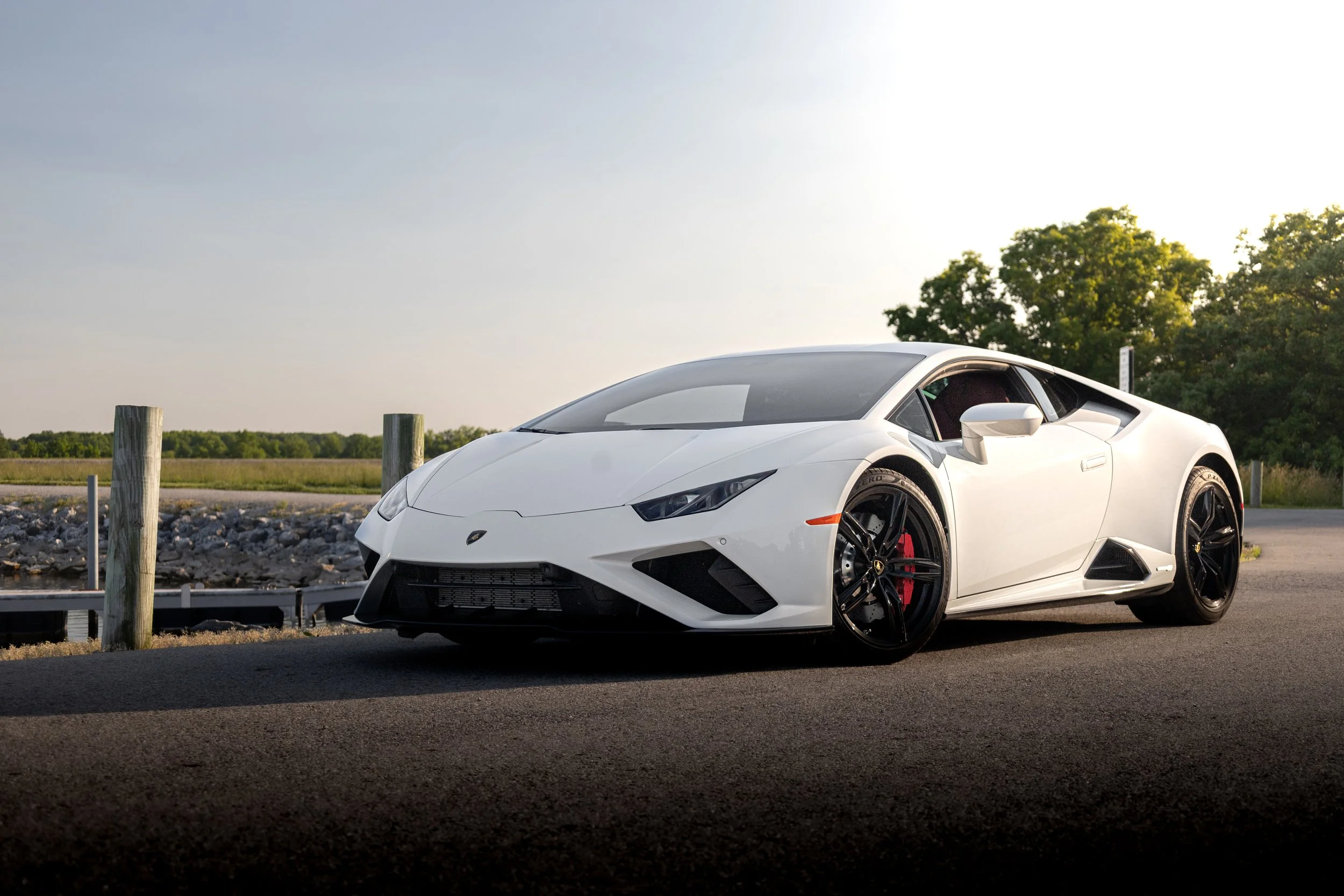 White Lamborghini sports car parked on a paved area near a body of water with trees in the background during daylight.