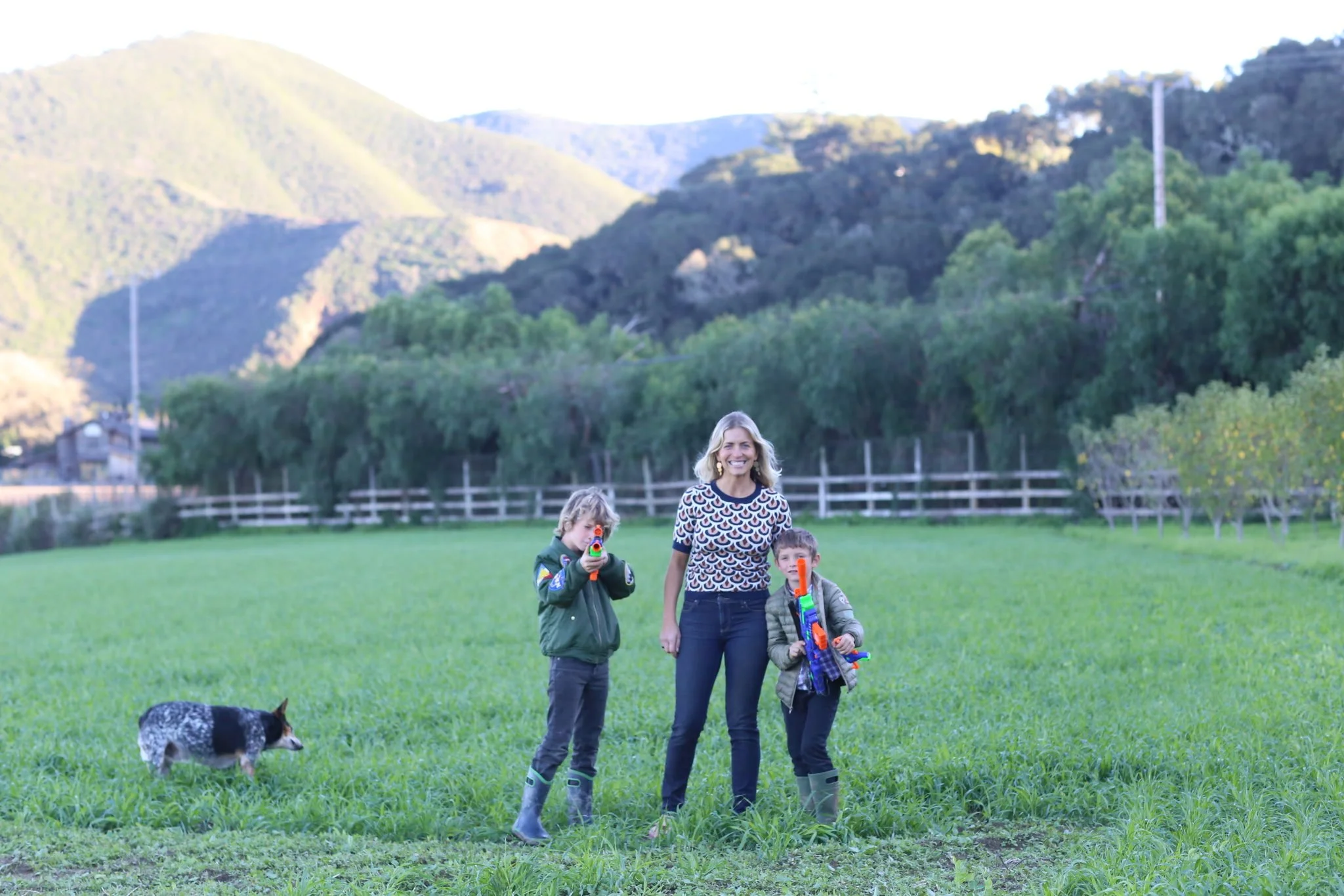 A woman and two children playing with toy guns in a grassy field, with a dog nearby, surrounded by mountains and trees in the background.