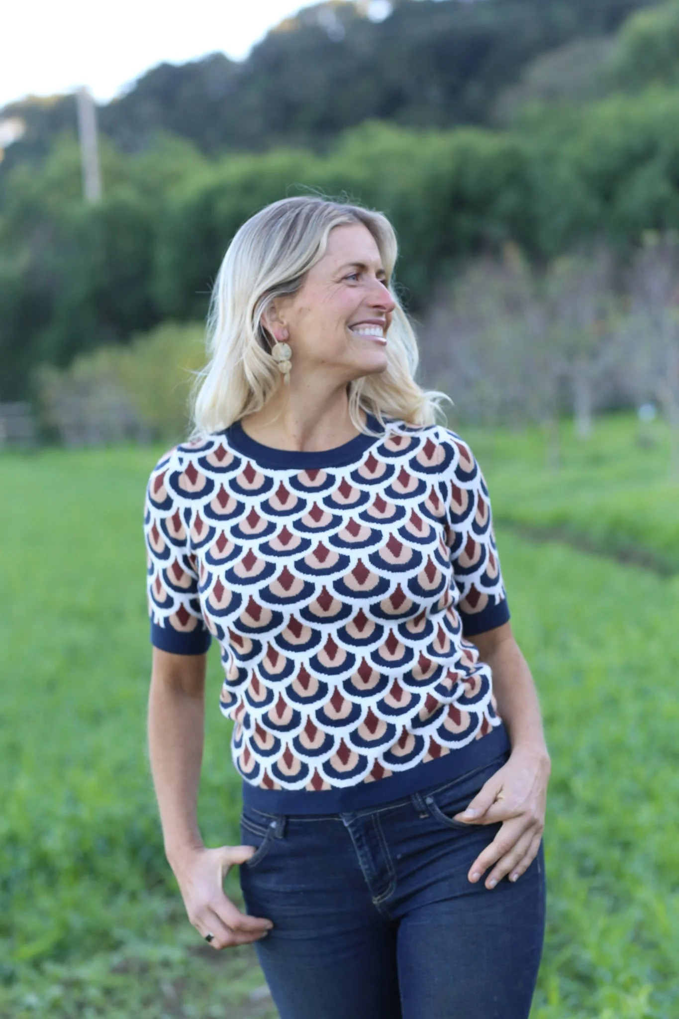 A woman with blonde hair smiling and looking to her right, standing outdoors in a lush green field with trees and hills in the background, wearing a patterned short-sleeved sweater and jeans.