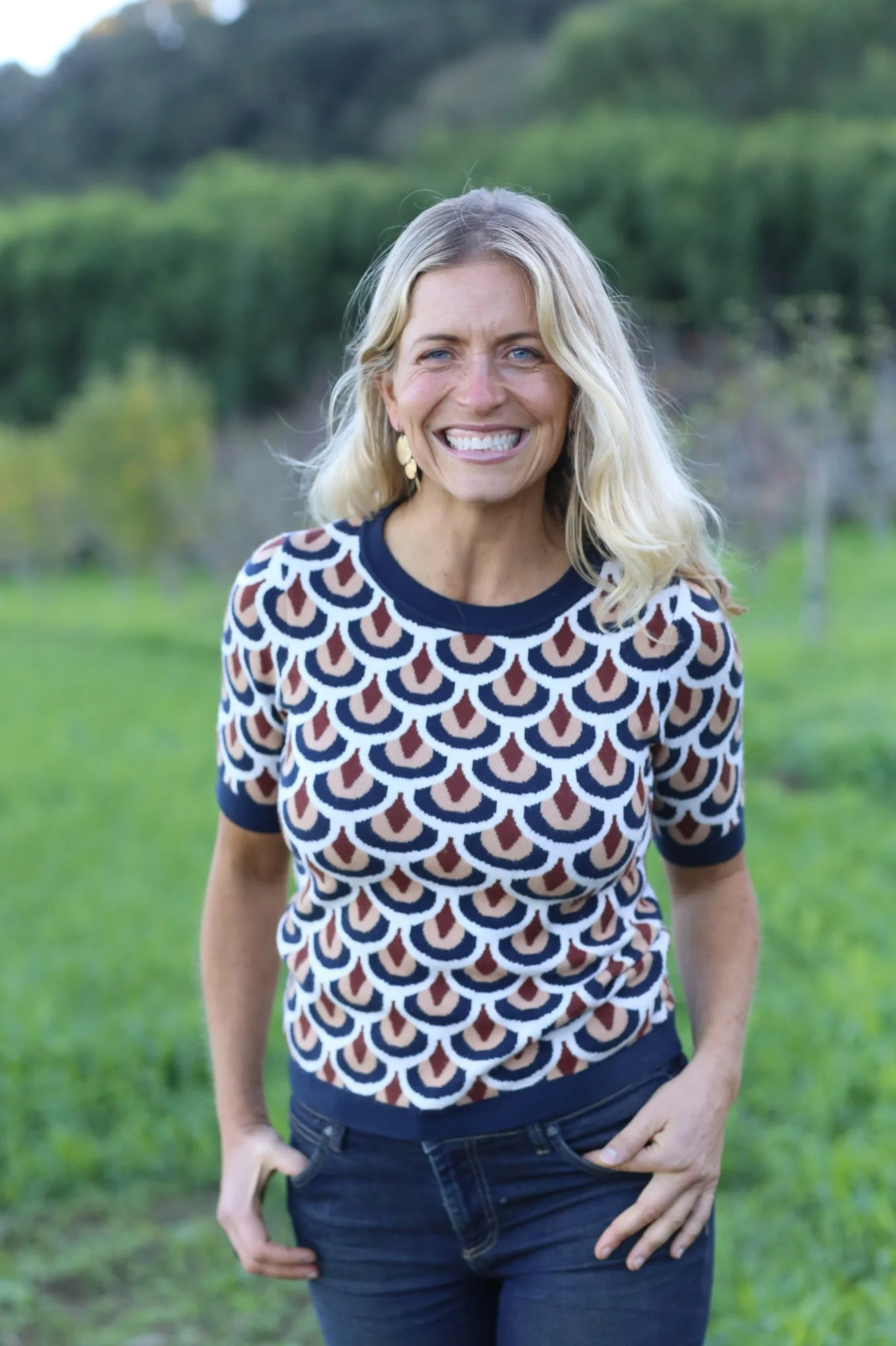 A smiling woman with blonde hair, wearing a patterned shirt with navy, brown, and white colors, standing outdoors on a grassy field with trees and hills in the background.