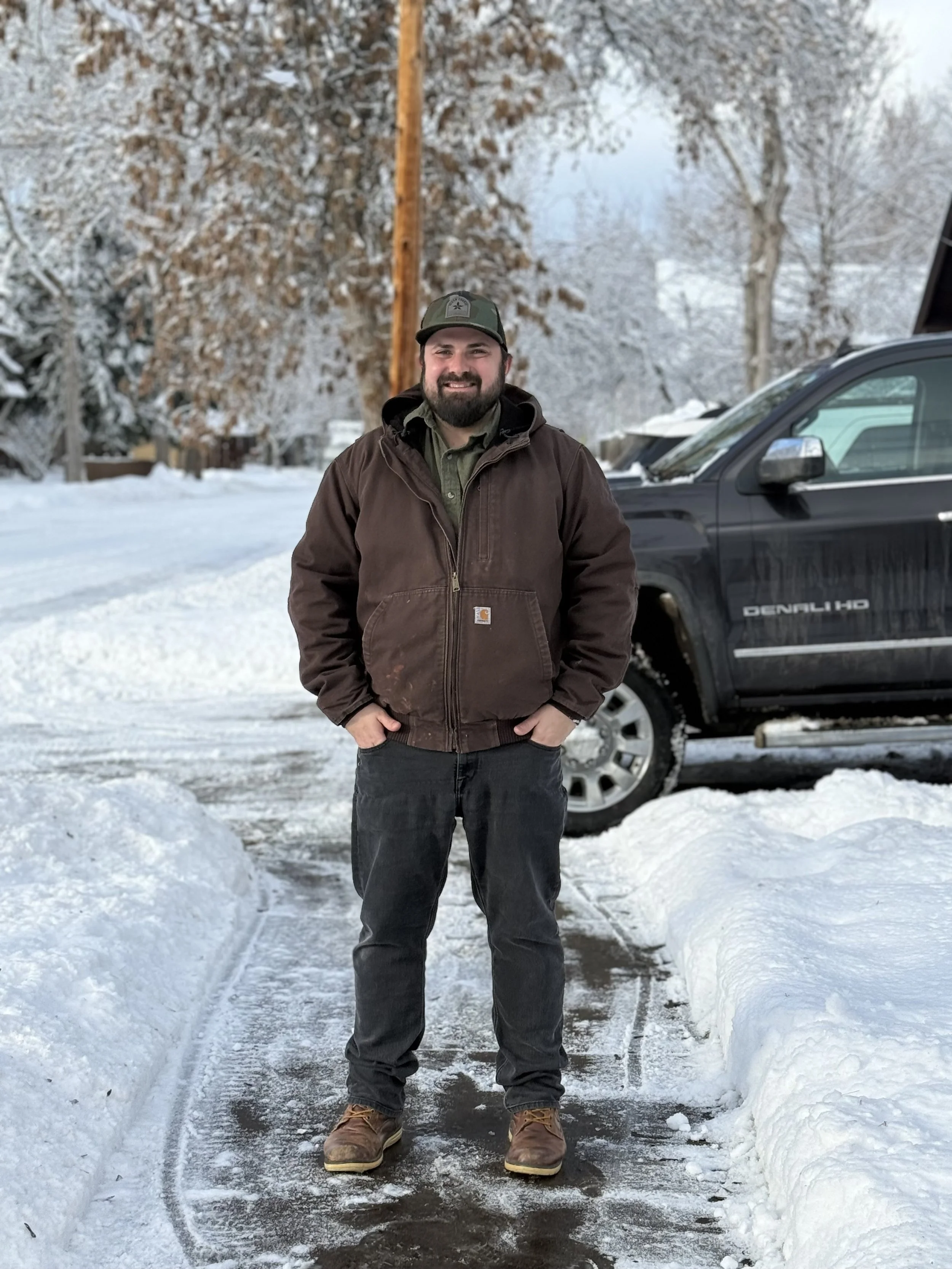 A man with a beard wearing a brown carhartt jacket, green shirt, cap, and brown boots stands on a snow-covered path with snow banks on either side, in front of a black vehicle, with snow-covered trees and a wooden utility pole in the background.