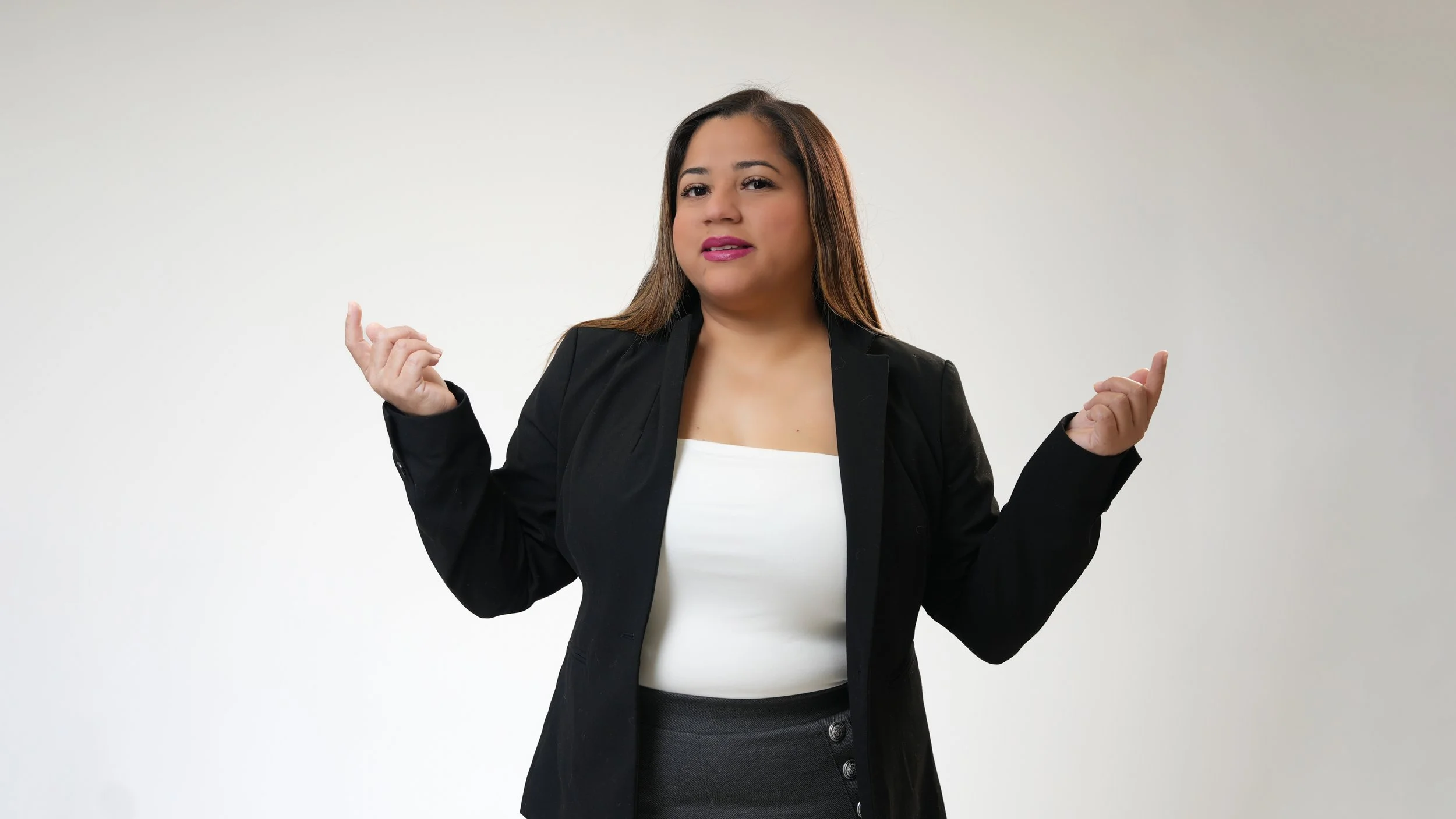 A woman with shoulder-length brown hair wearing a black blazer, white top, and dark skirt, standing against a plain white background with her arms slightly raised and fingers relaxed.