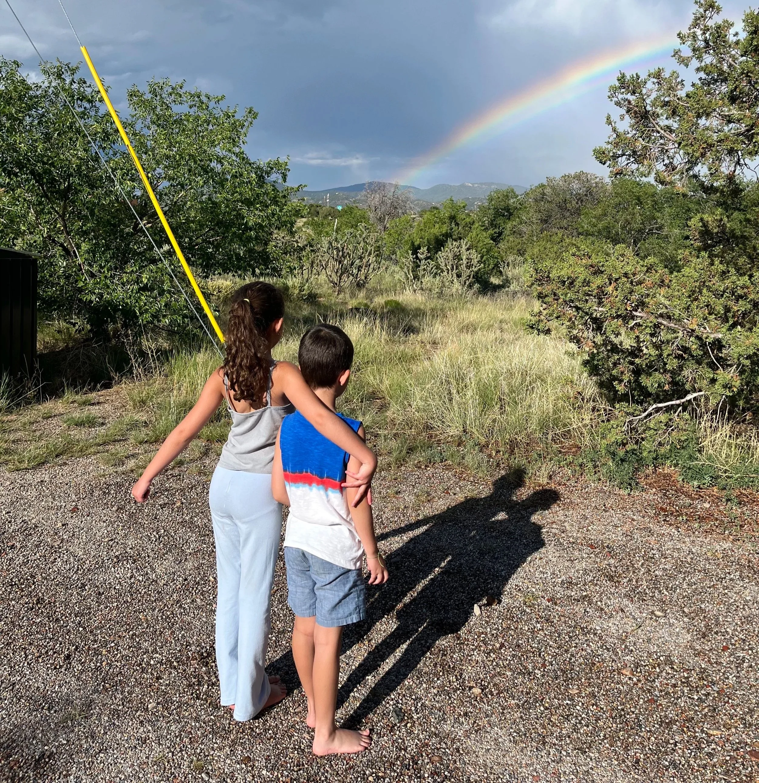 Two children, a girl and a boy, standing on a gravel path watching a rainbow in the sky over greenery and mountains in the distance during a rainstorm.