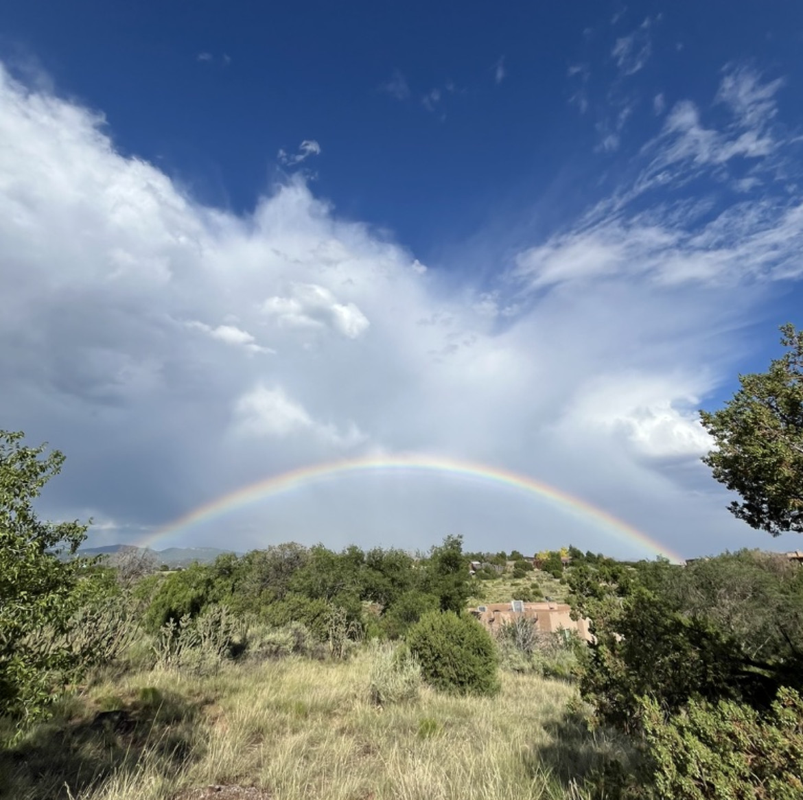 A landscape scene with a rainbow arching across a partly cloudy sky over green hills with trees and houses.