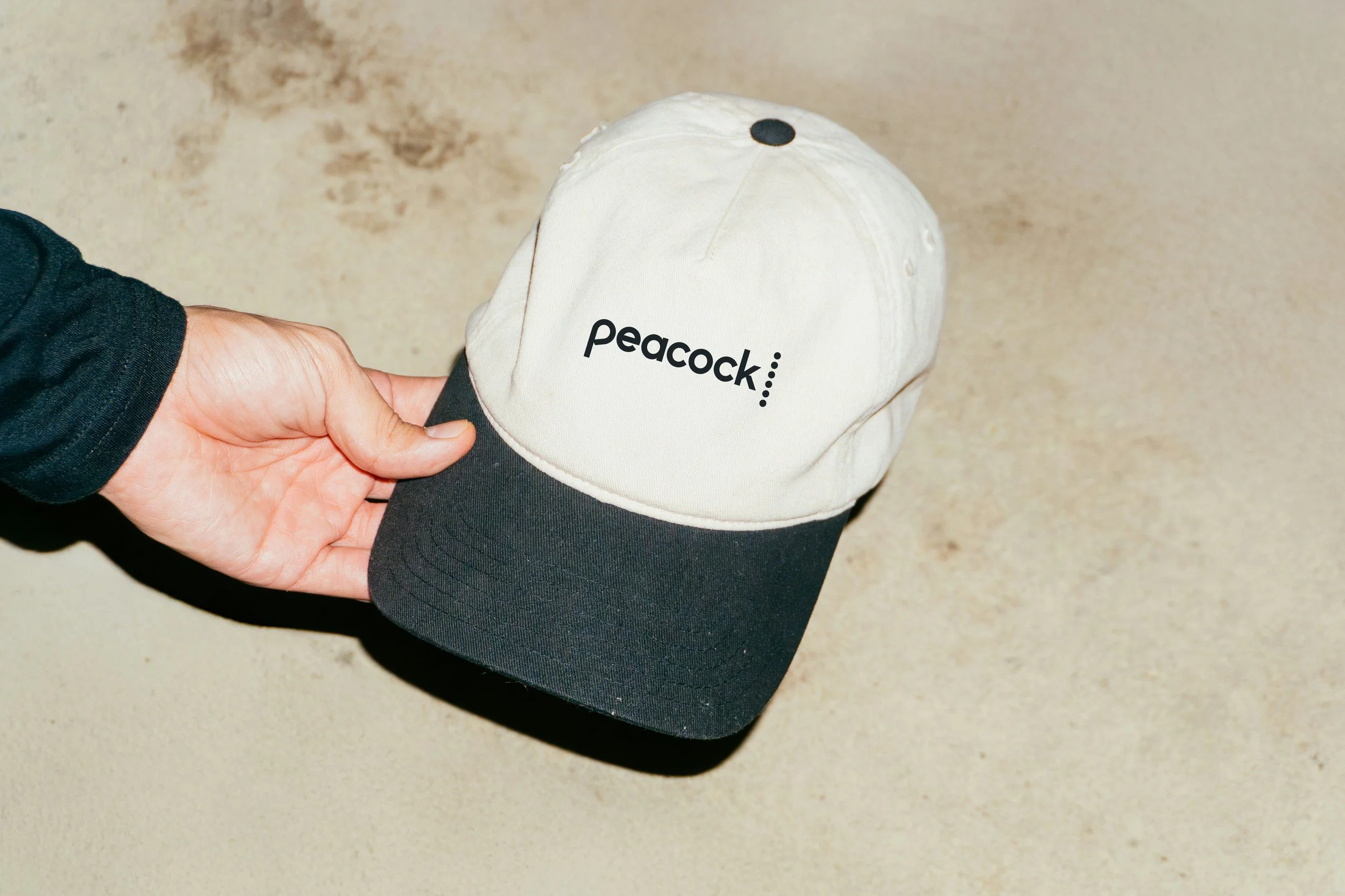Person holding a beige and black baseball cap with 'peacock' embroidered on the front, against a beige textured background.