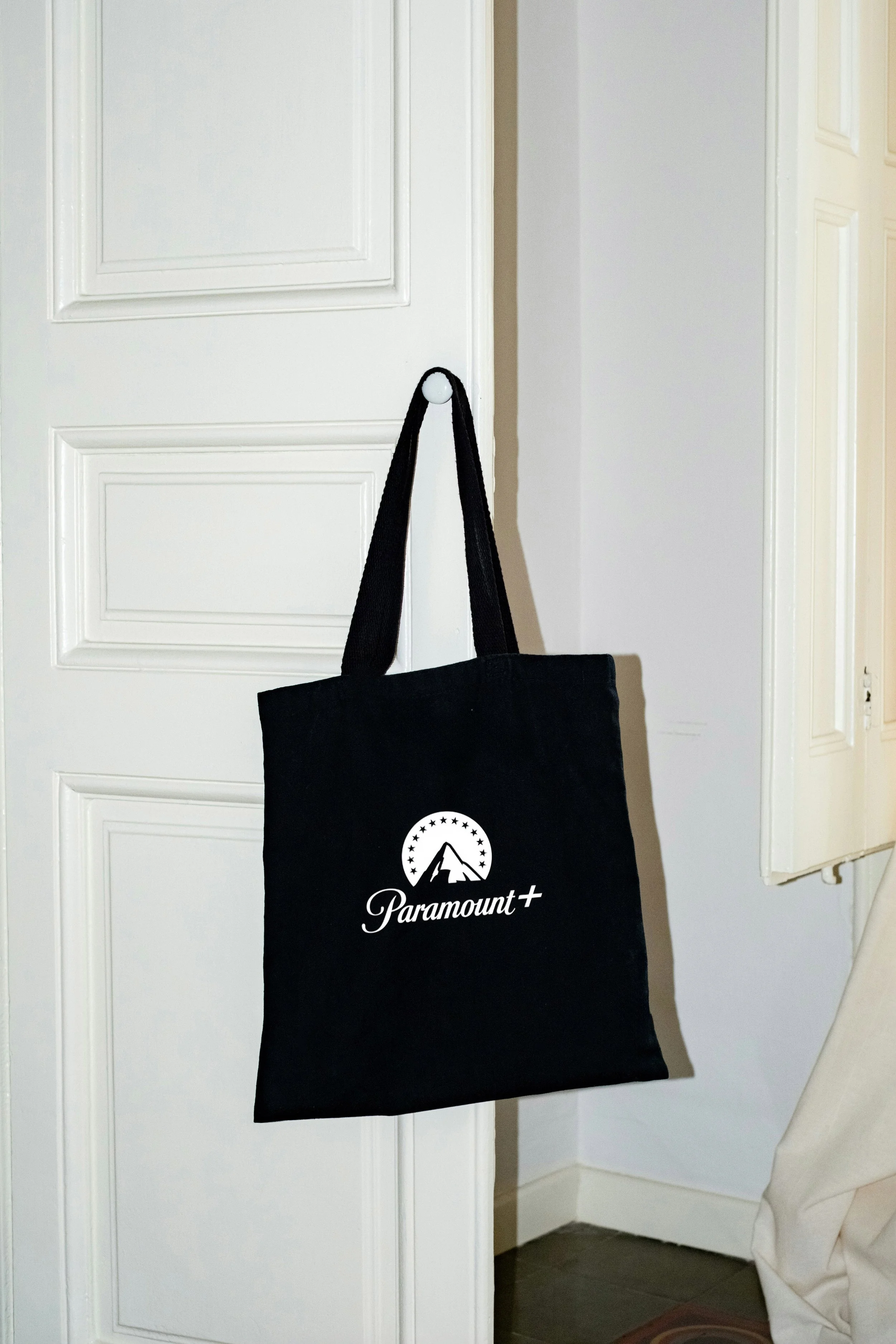 Black tote bag hanging on a white door, with a white Paramount+ logo featuring a mountain and stars.